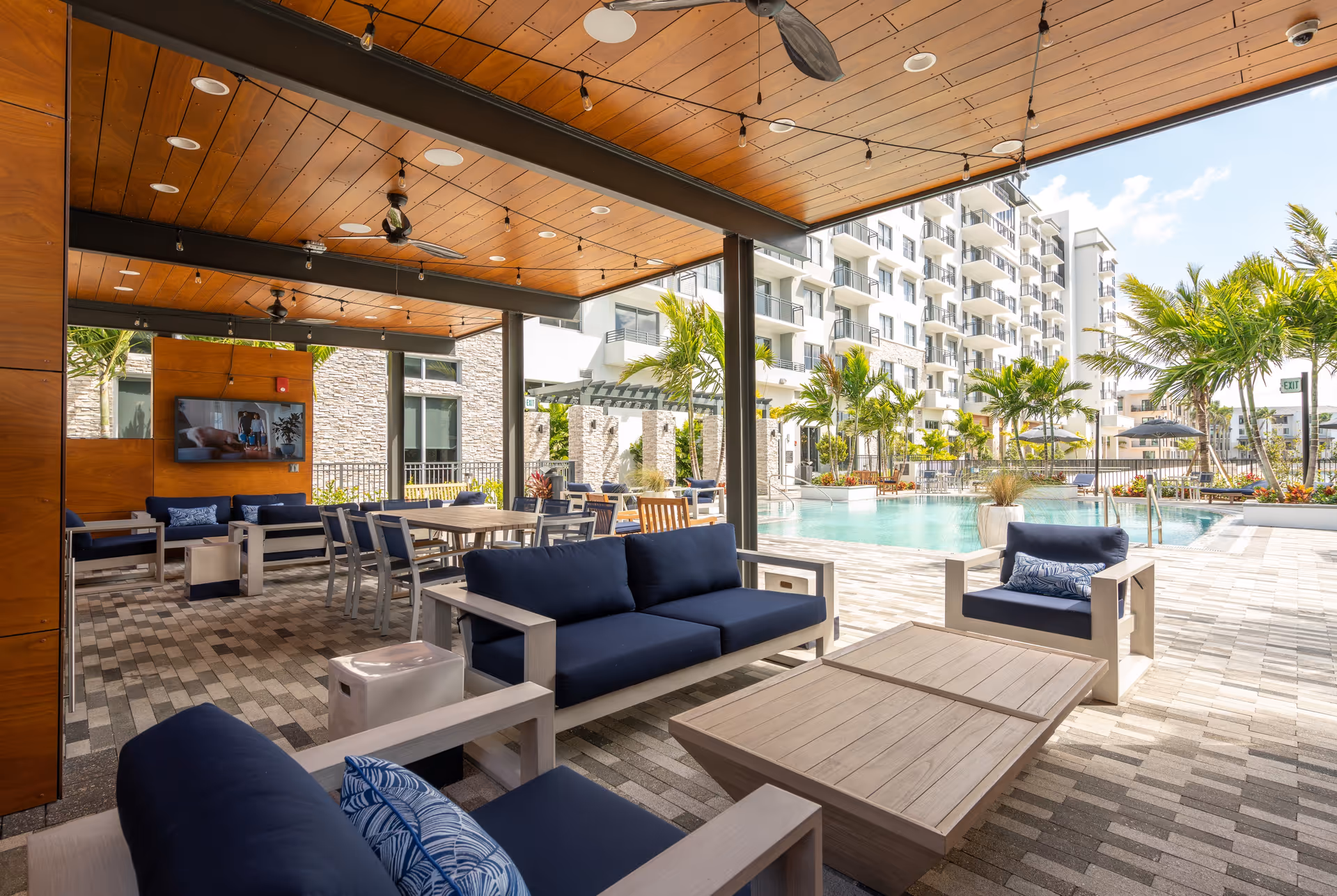 Covered outdoor lounge area with navy cushioned seating and dining tables beside a swimming pool and a multi-story residential building.