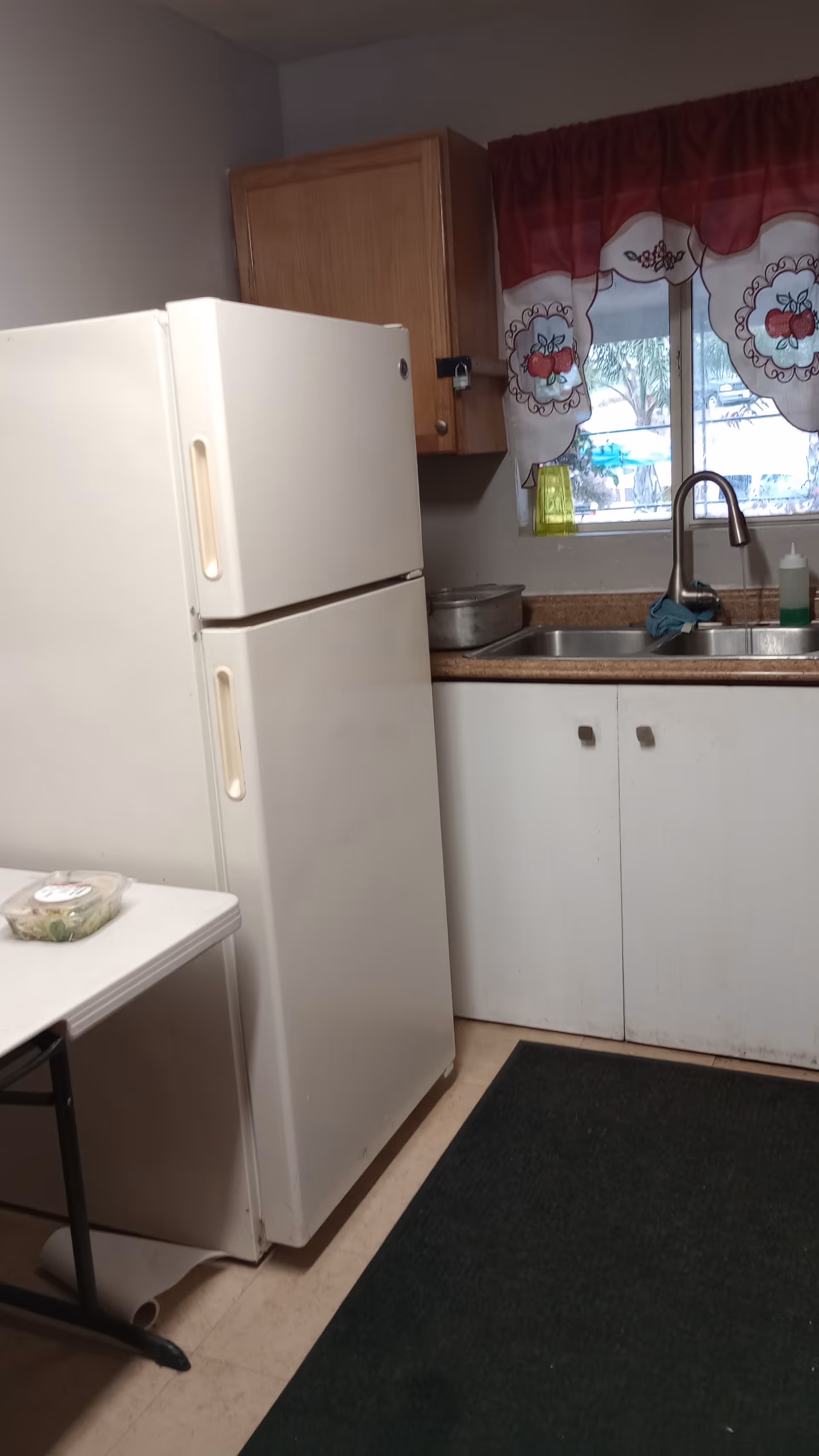 Compact kitchen with a refrigerator, sink, cabinets, and a window with decorative curtains.