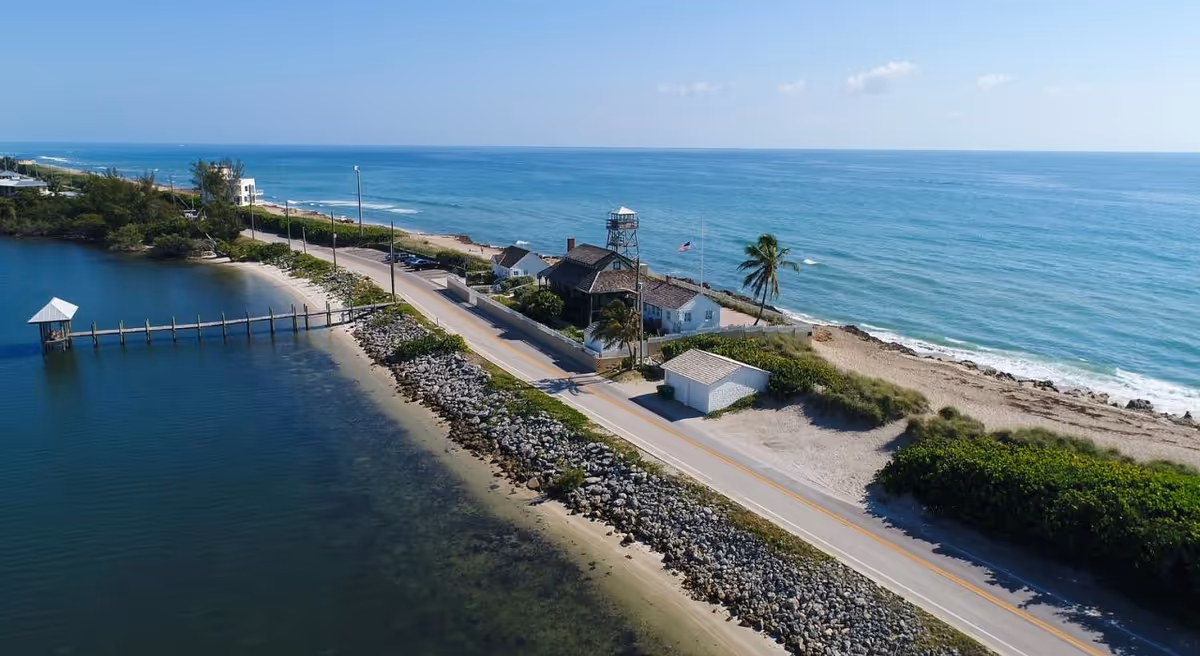 Aerial coastal scene with a road, beach, ocean, and small buildings along the shore.