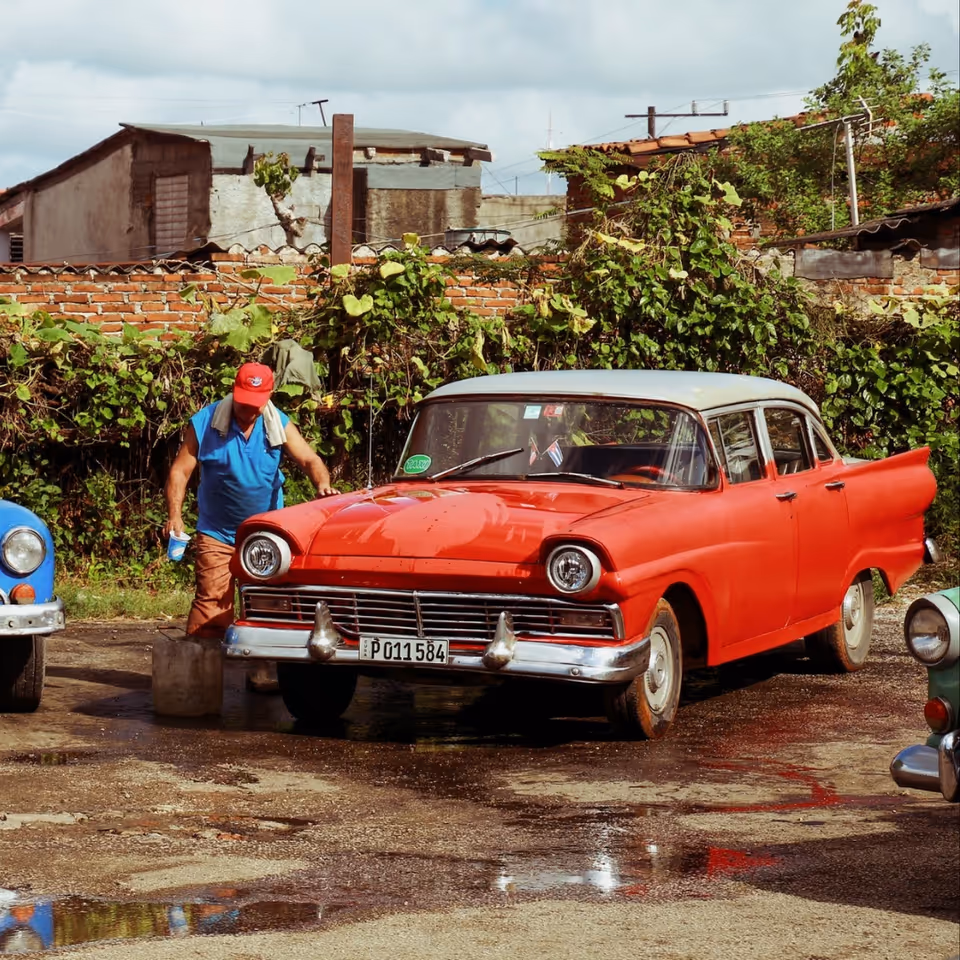 A man in a blue shirt and red cap cleans a vintage red car outdoors.