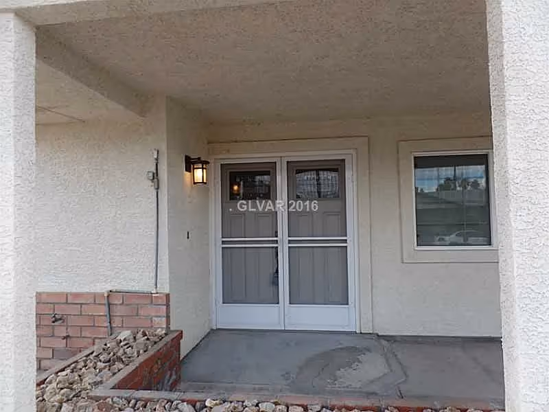 Covered front porch showing double glass entry doors, a wall lantern, and a small rock garden at a stucco house entrance.