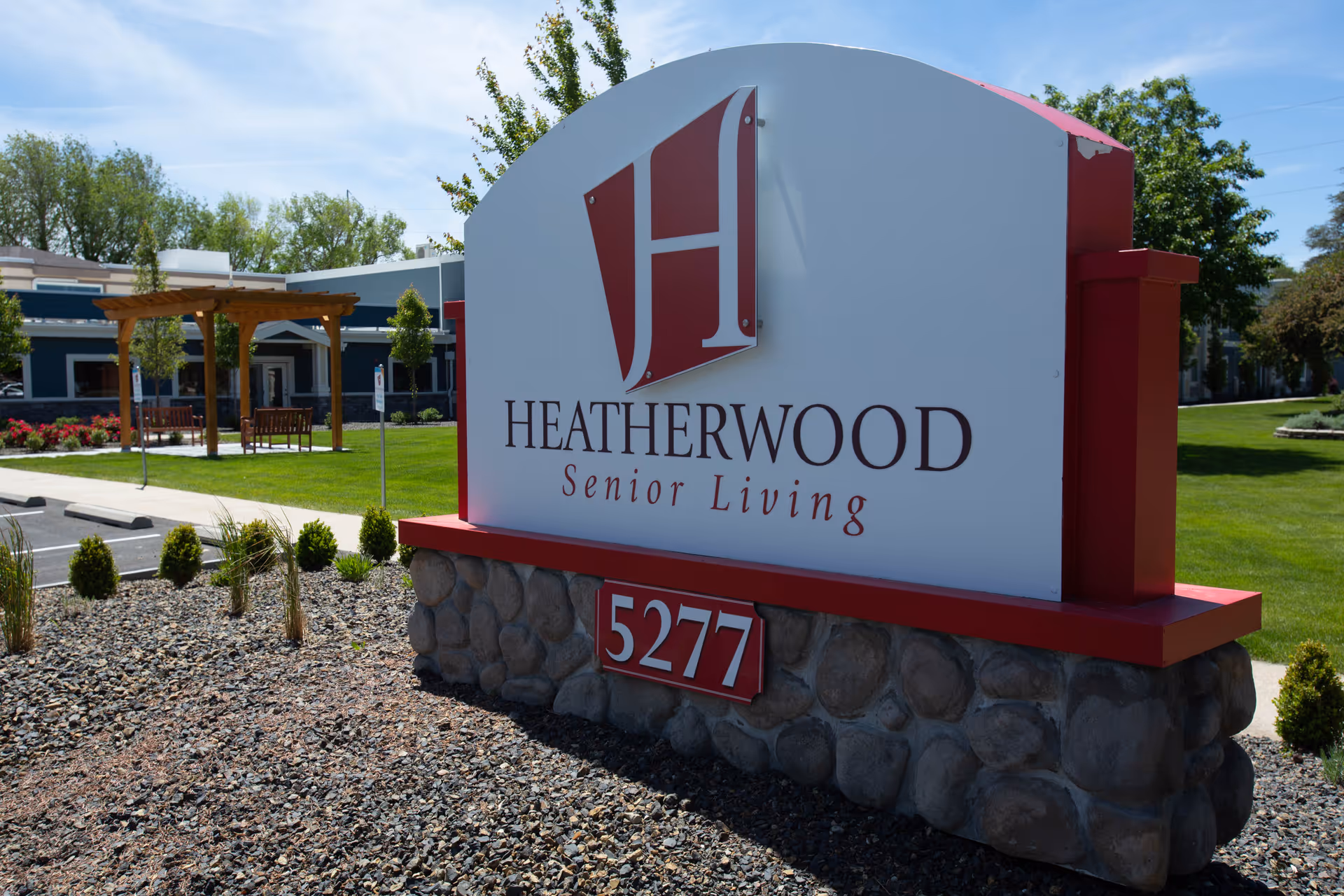 Outdoor view of the Heatherwood Senior Living facility sign with the address number 5277. The sign is mounted on a stone base with a landscaped area of rocks and small bushes around it. In the background, there is a building with a pergola, benches, green grass, and trees under a clear sky.