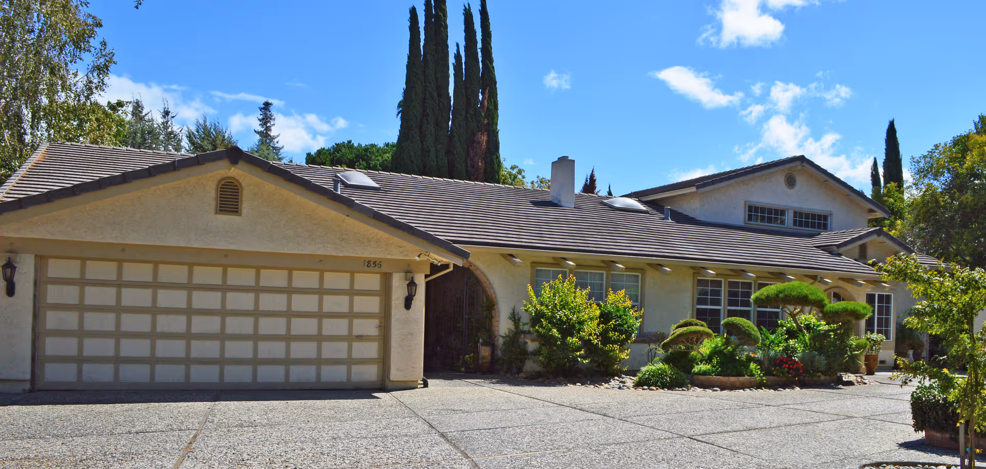 Exterior view of a single-story residential building with a two-car garage, beige stucco walls, and a tiled roof. The driveway is paved with textured concrete, and there are well-maintained shrubs and trees in front of the house under a blue sky with some clouds.