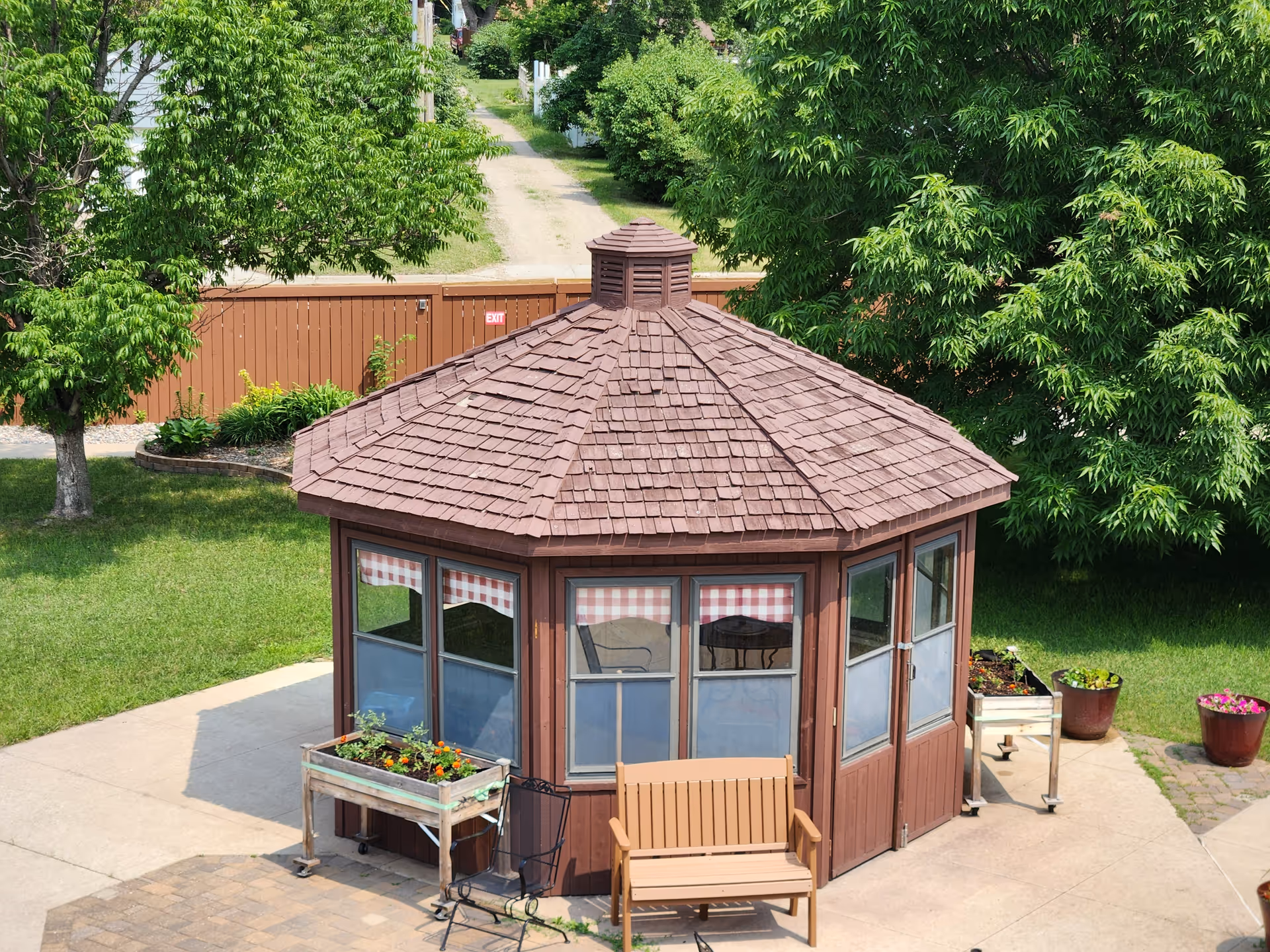 Brown hexagonal gazebo in a courtyard with benches, planters, and green trees.