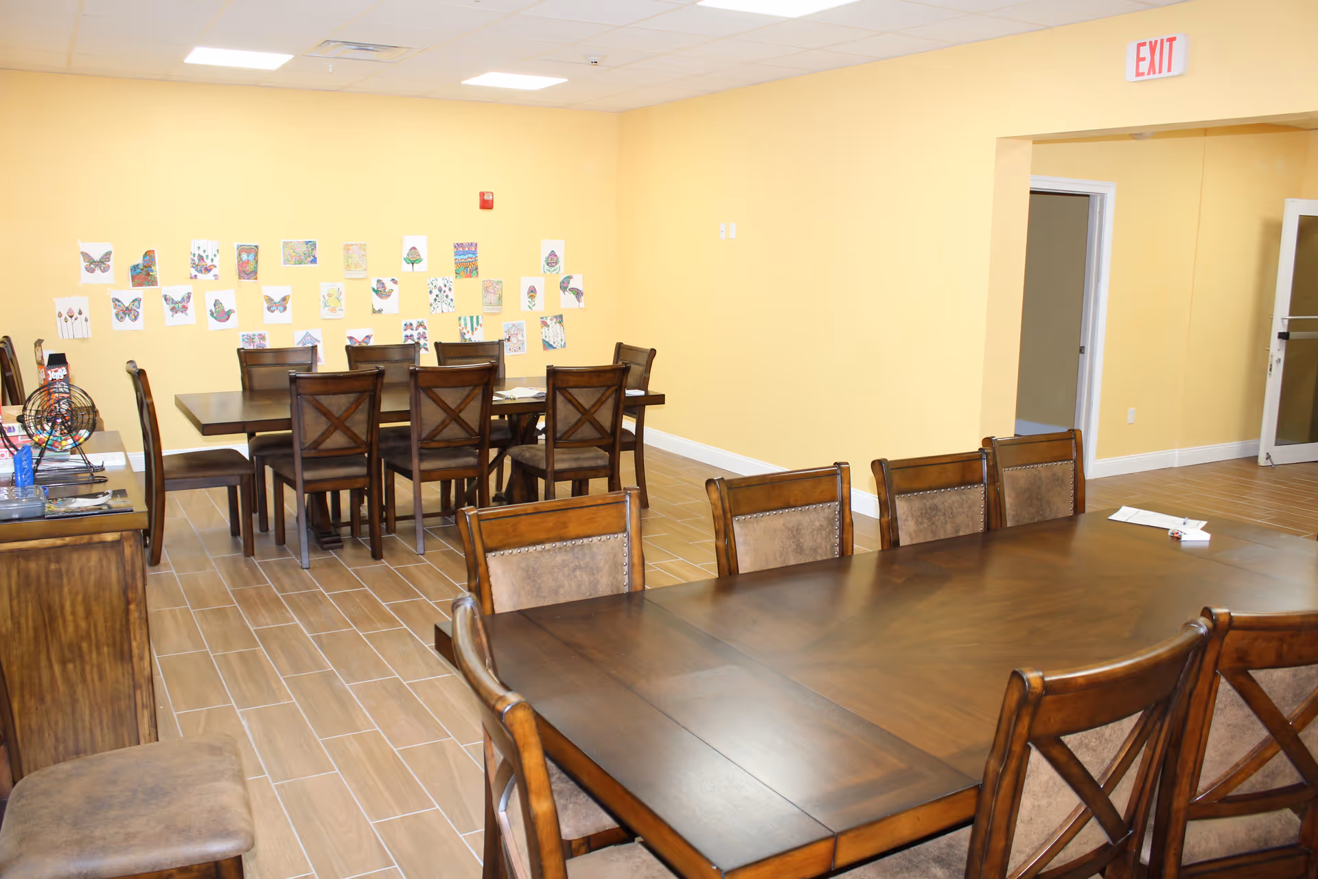 Bright dining room with long wooden tables, chairs, and colorful wall art in a care facility.