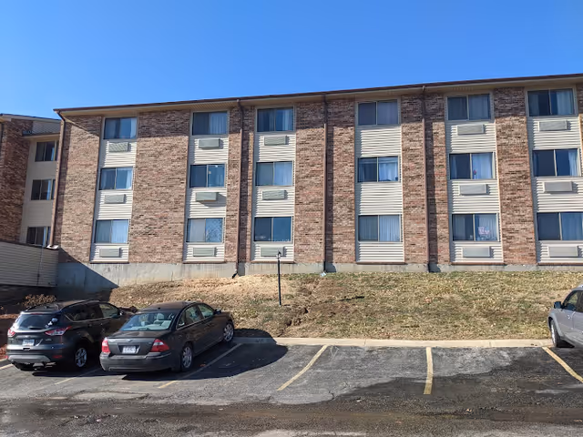 Exterior view of a three-story brick building with multiple windows, some with air conditioning units. In front of the building, there is a small grassy area and a parking lot with three parked cars under a clear blue sky.