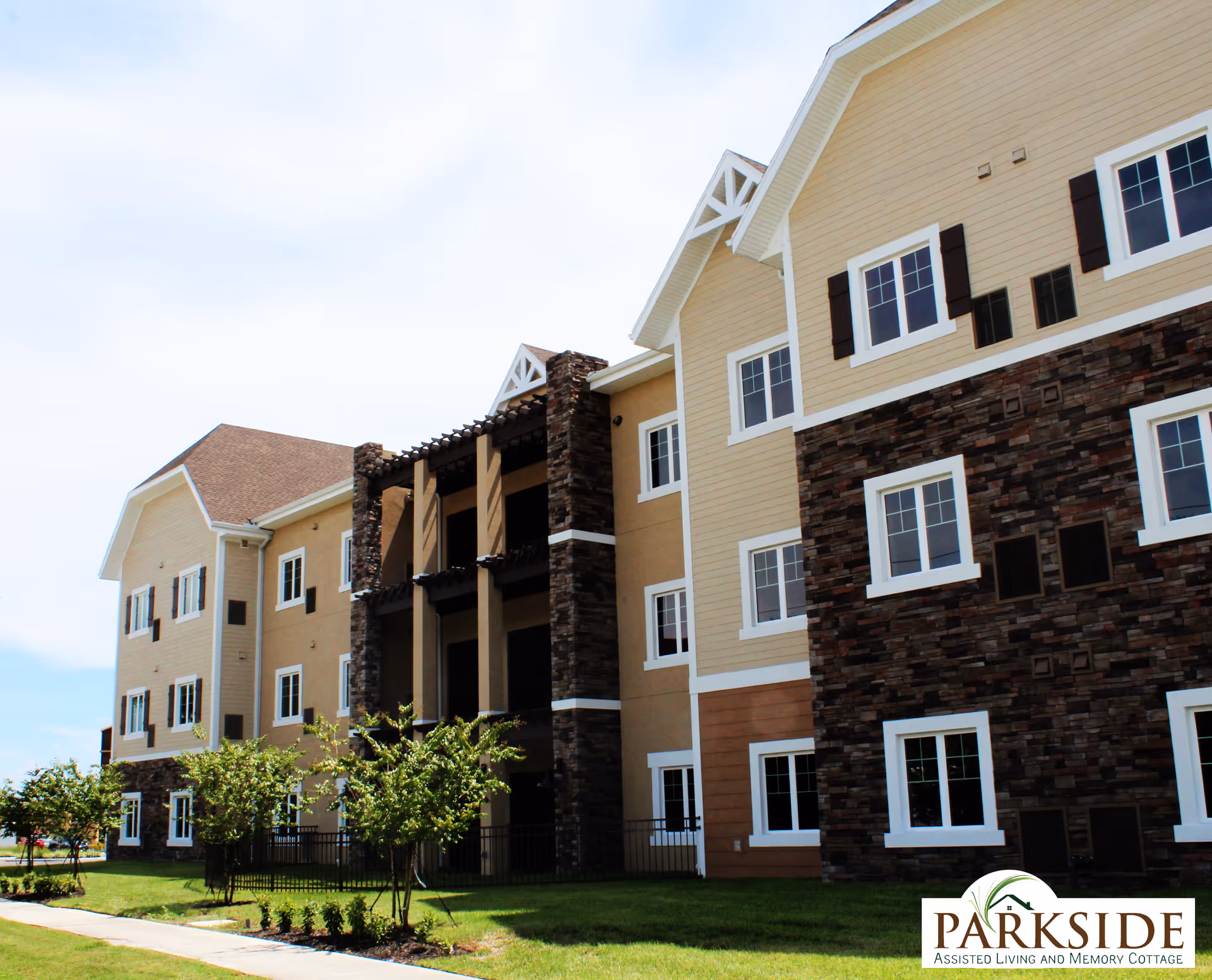 Exterior view of a multi-story assisted living facility building with beige siding and stone accents, surrounded by a well-maintained lawn and small trees under a partly cloudy sky.