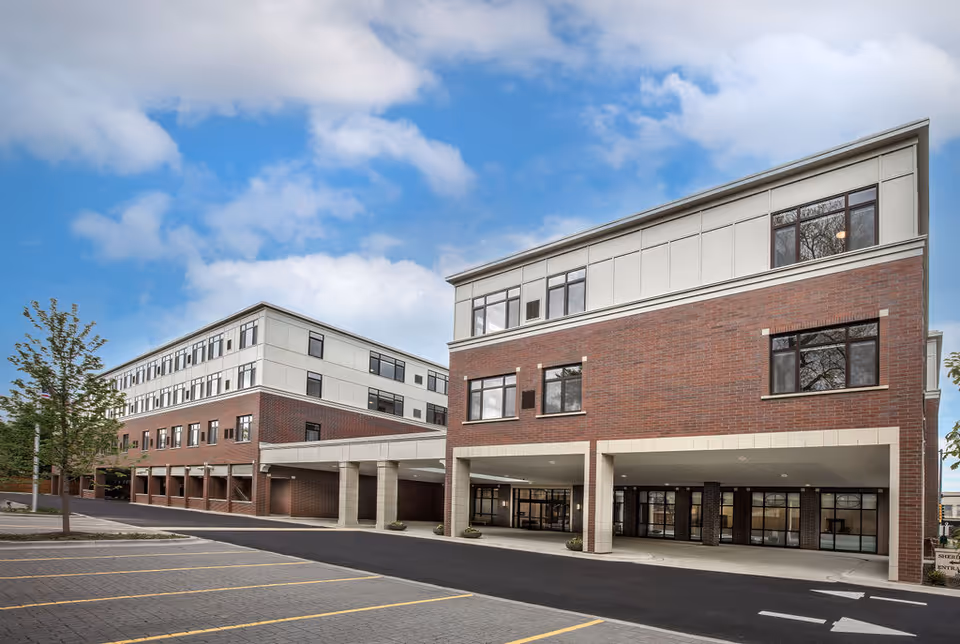 Exterior view of a modern multi-story senior living facility building with brick and light-colored paneling under a partly cloudy blue sky. The building has large windows and a covered entrance area with columns. There is a parking lot with marked spaces in the foreground and a small tree on the left side.