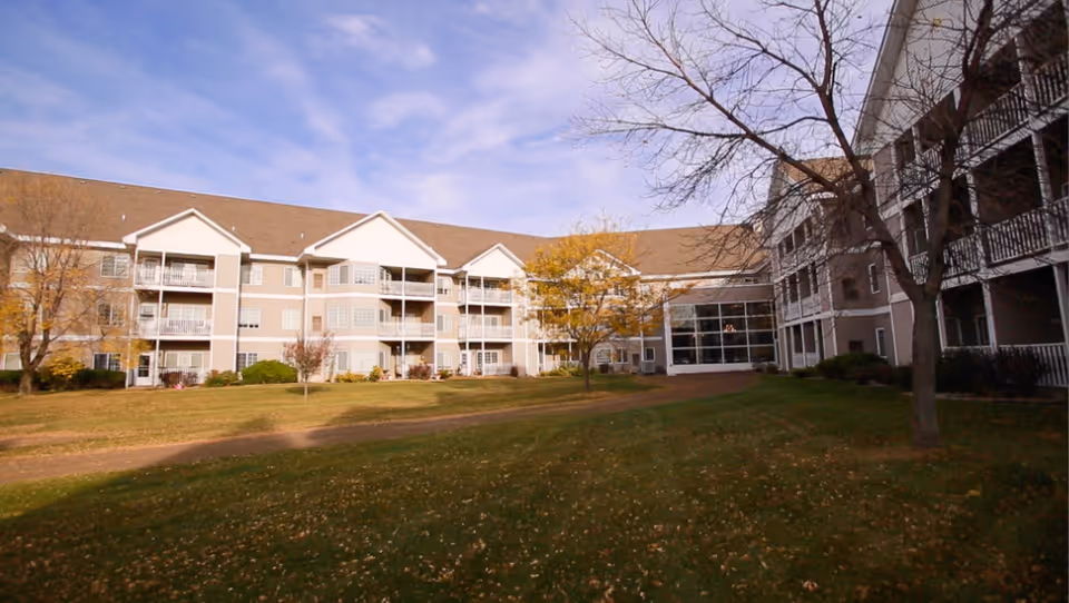 Exterior view of a three-story senior living building with balconies overlooking a grassy courtyard and trees.