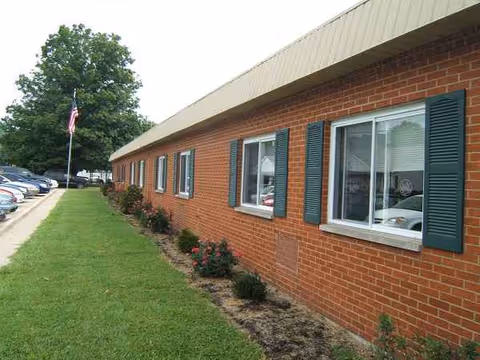 Brick nursing home exterior with green shutters along a grassy walkway and parked cars.