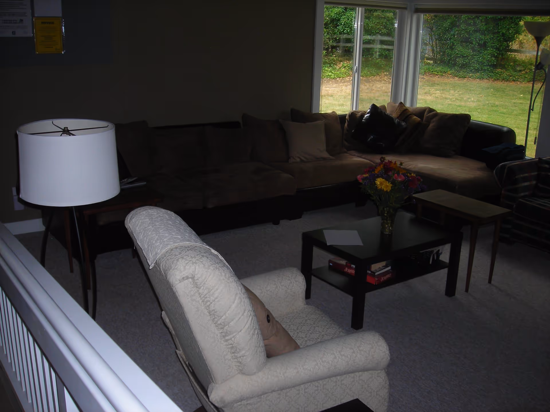 Cozy living room with a sectional sofa, upholstered armchair, coffee table with flowers and a floor lamp in front of a large window showing a grassy yard.