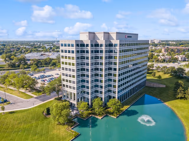 Tall glass-front office building alongside a turquoise pond and green lawn.
