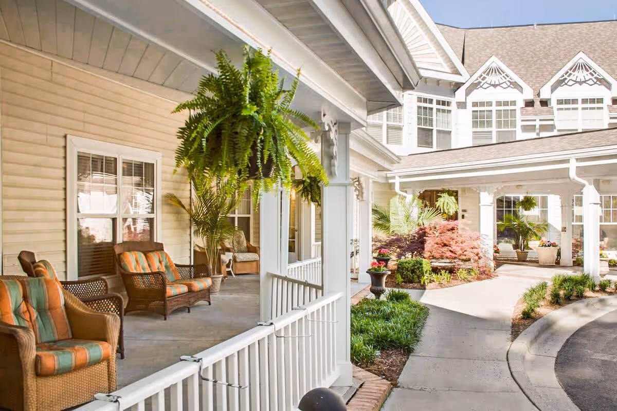 Covered porch with wicker seating, hanging ferns, and a landscaped entrance to a senior living building.