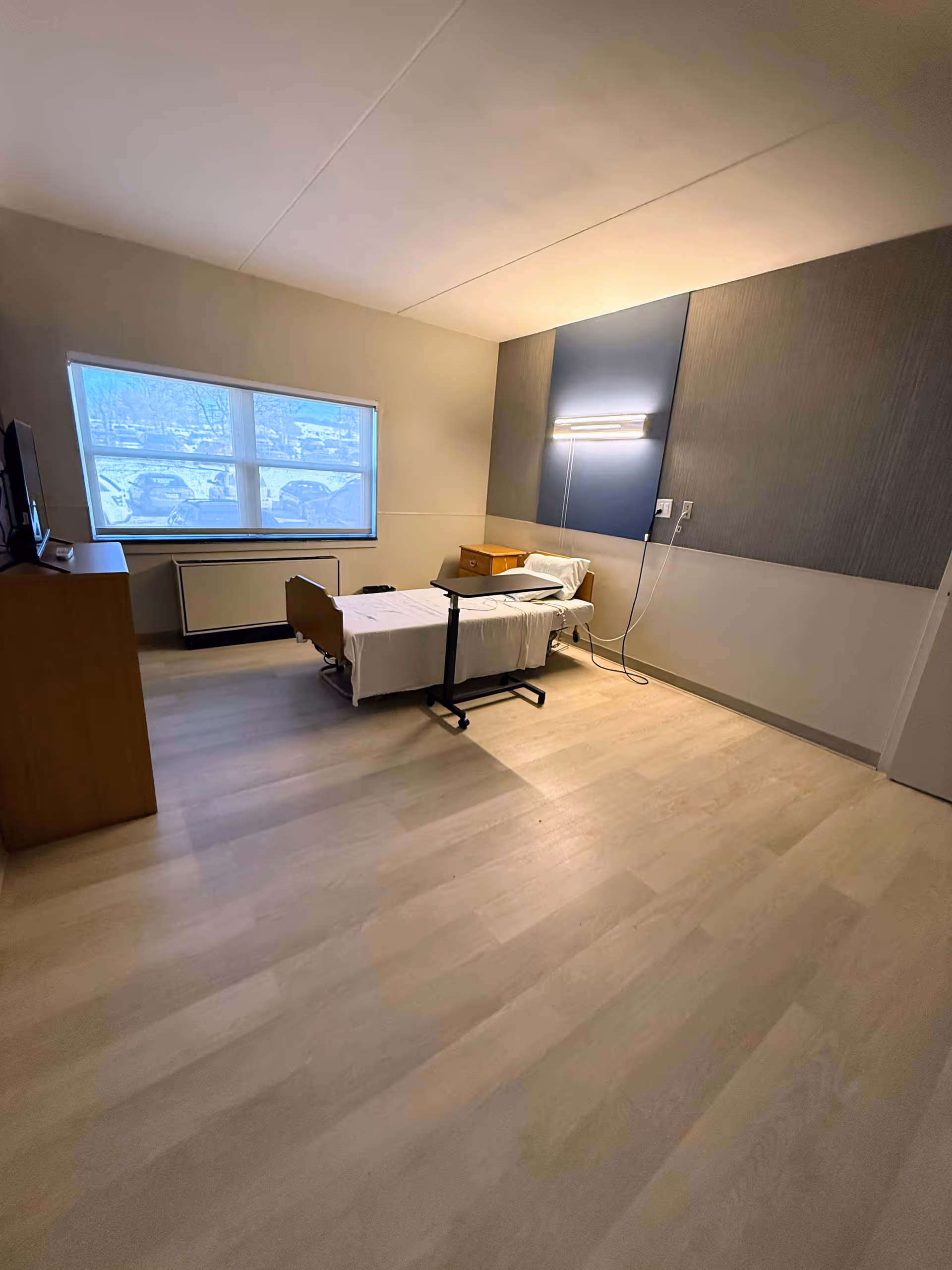 Single occupancy patient bedroom with bed, dresser, window, and soft-blue wall panels.