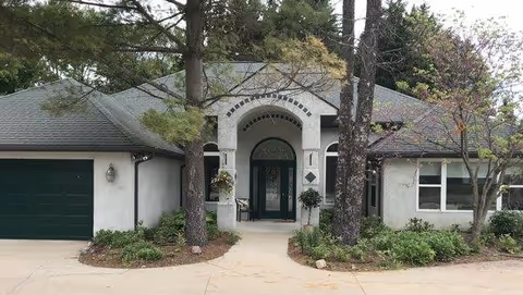 A single-story care home exterior with an arched entrance, green door, and surrounding shrubs.