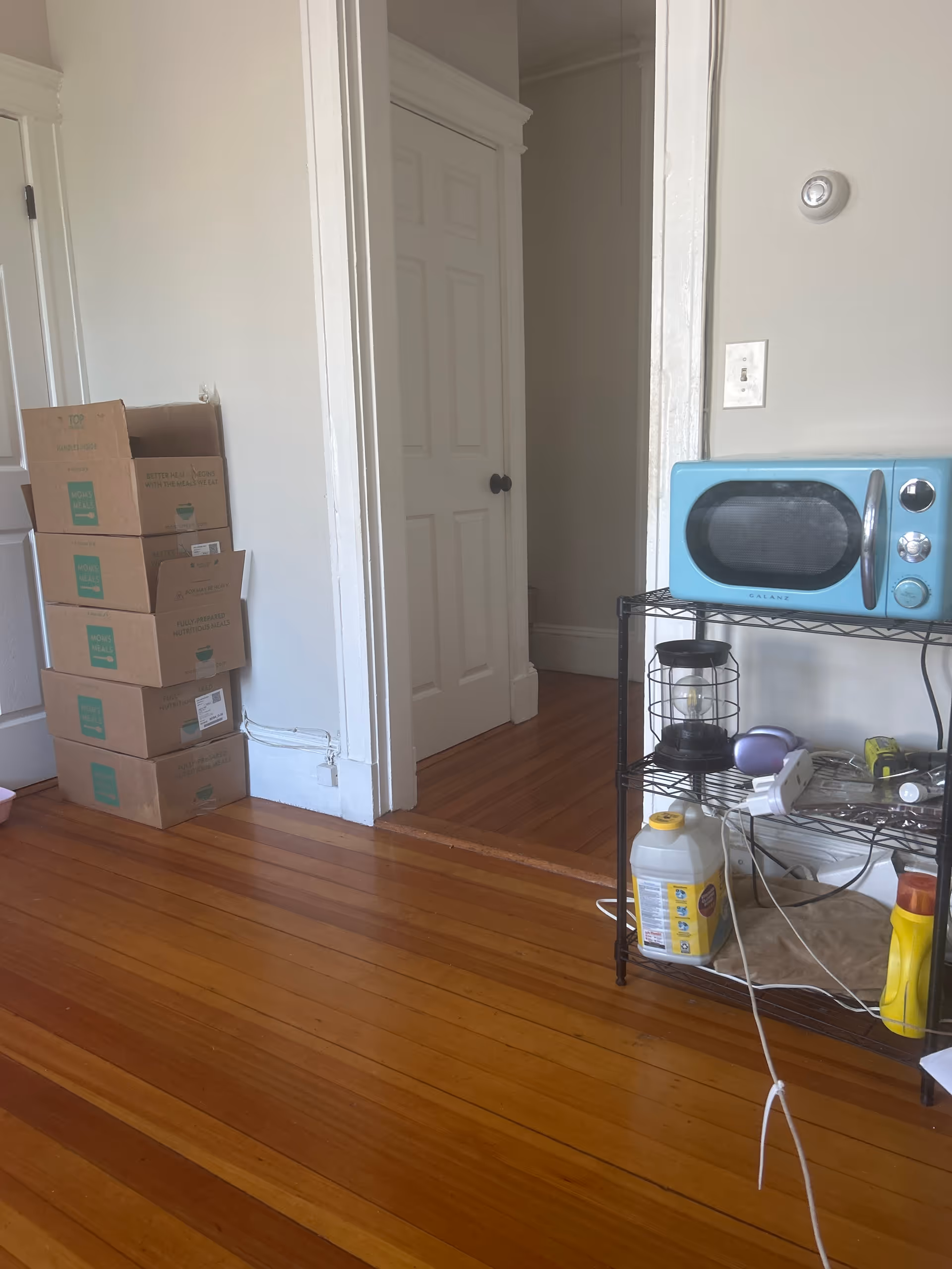 Interior living area with a blue microwave on a metal rack, stacked boxes, and wooden floors.