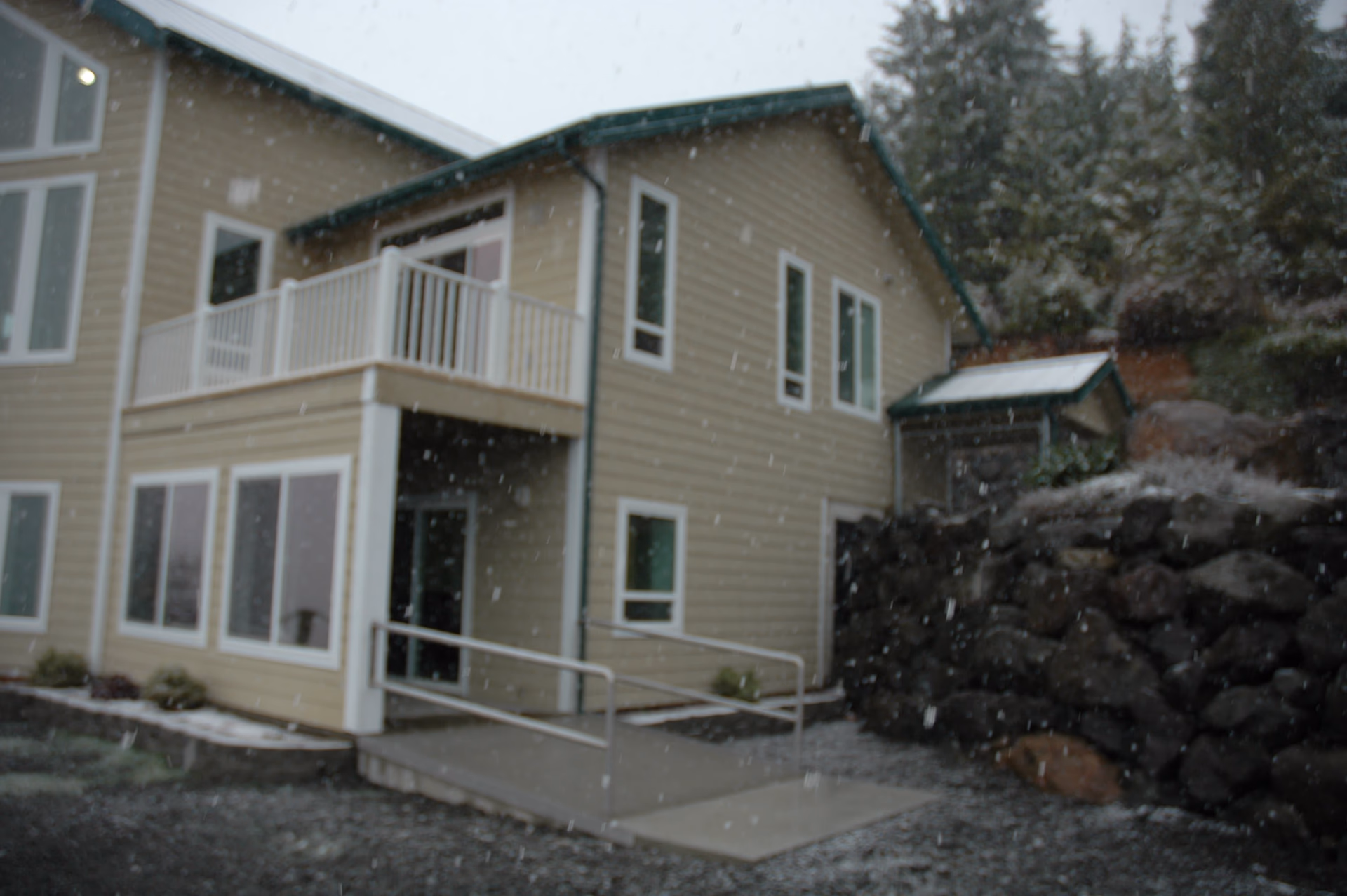 Exterior view of a two-story beige building with white trim and a green roof, featuring a balcony and a wheelchair-accessible ramp. Snow is falling, and there are trees and rocks surrounding the building.