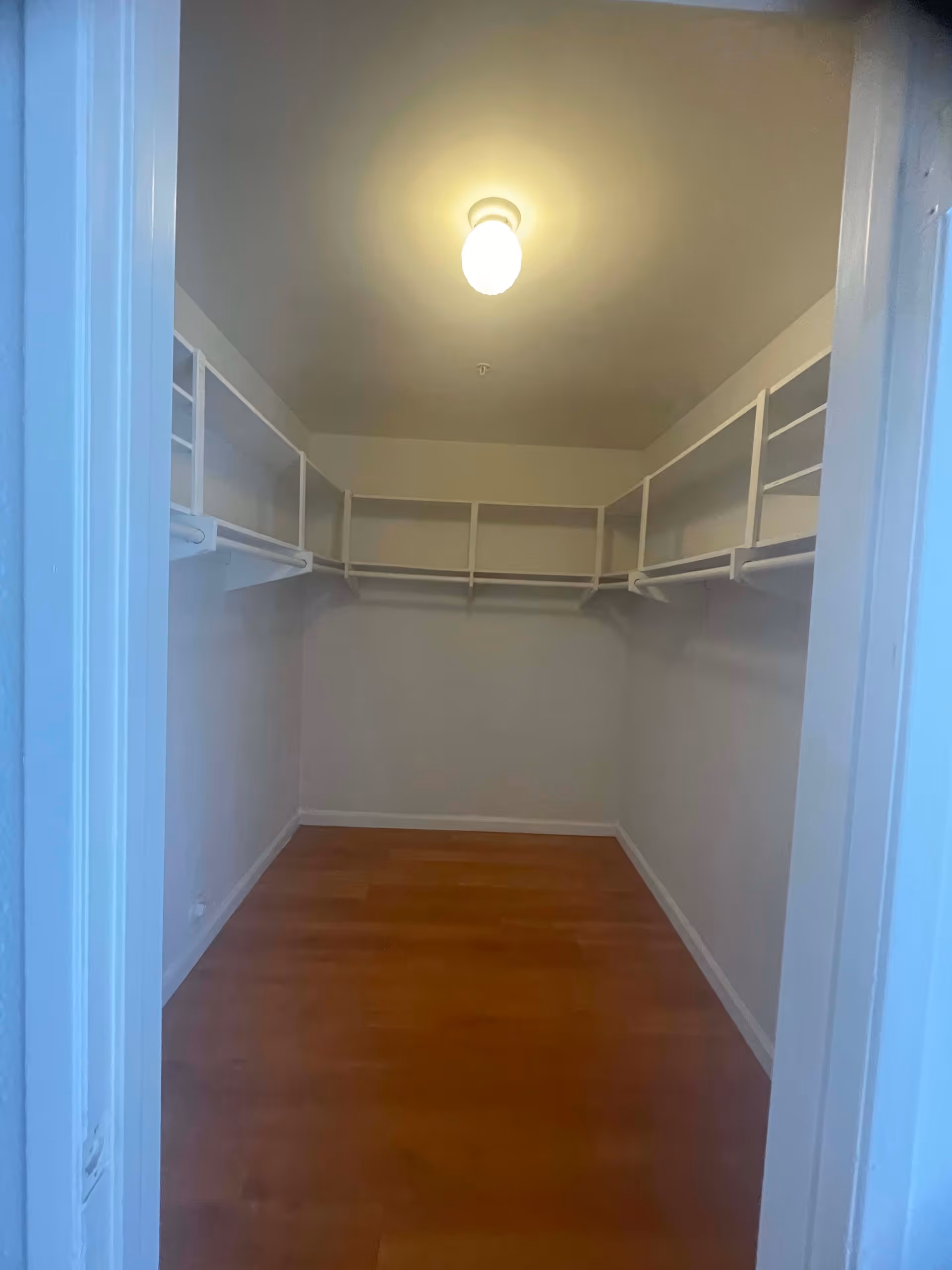 Empty walk-in closet with wooden floor, white walls, and white shelving units along the upper perimeter. A single ceiling light fixture is turned on.
