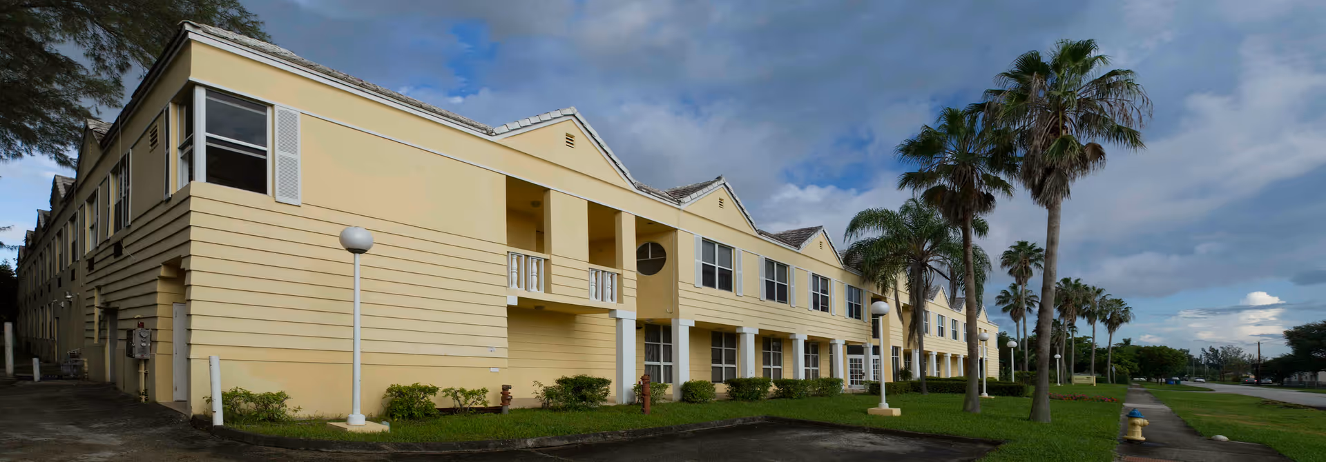 Exterior view of a two-story yellow building with white trim and multiple windows, surrounded by palm trees and a grassy area with a sidewalk and street lamps under a partly cloudy sky.