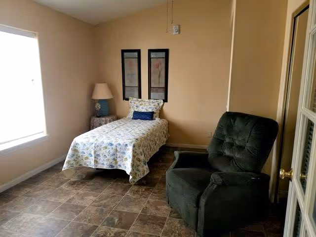 A small bedroom with a single bed covered in a floral bedspread and a blue pillow. Next to the bed is a nightstand with a lamp. There are two framed artworks hanging on the wall above the bed. A dark green upholstered armchair is positioned near the door. The room has a large window letting in natural light and a tiled floor.