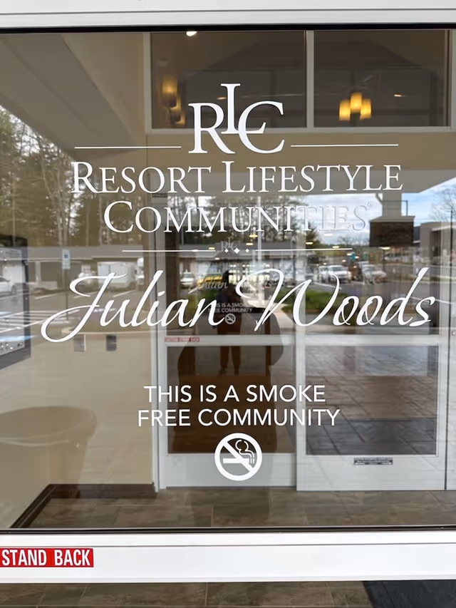 Glass entrance door of Julian Woods Retirement Community with white text displaying the name 'Resort Lifestyle Communities Julian Woods' and a notice stating 'This is a smoke free community' with a no smoking symbol. The reflection shows part of the parking lot and surrounding area outside.