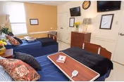 Two beds in a bright senior living bedroom with dresser, TV, clock, and wall art.
