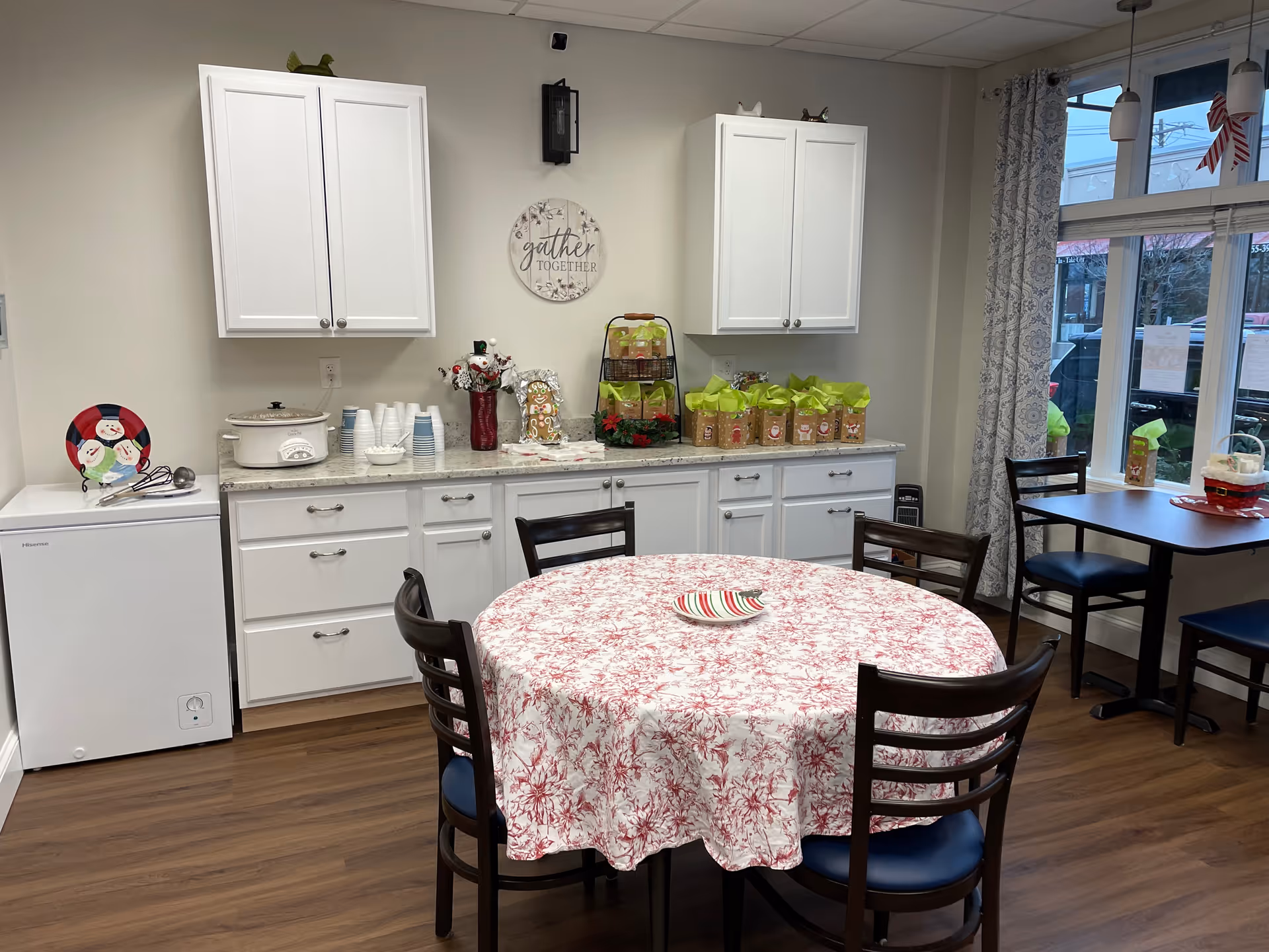 Communal dining area with round table, white cabinets, snacks, and a windowed seating nook.