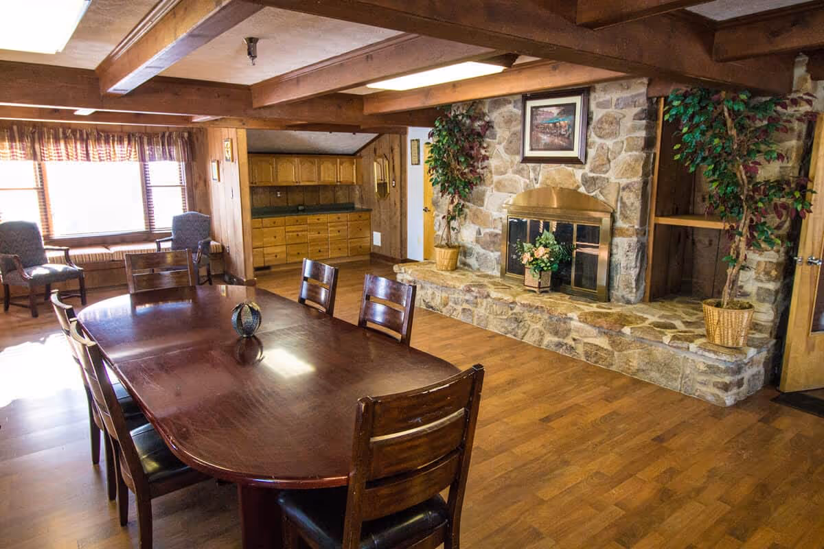 A cozy dining room with a large wooden table surrounded by six chairs. The room features a stone fireplace with a brass screen, two potted plants on either side, and a framed picture above the fireplace. There are two upholstered chairs near a window with plaid curtains, wooden floors, and exposed wooden beams on the ceiling. A built-in wooden cabinet is visible in the background.