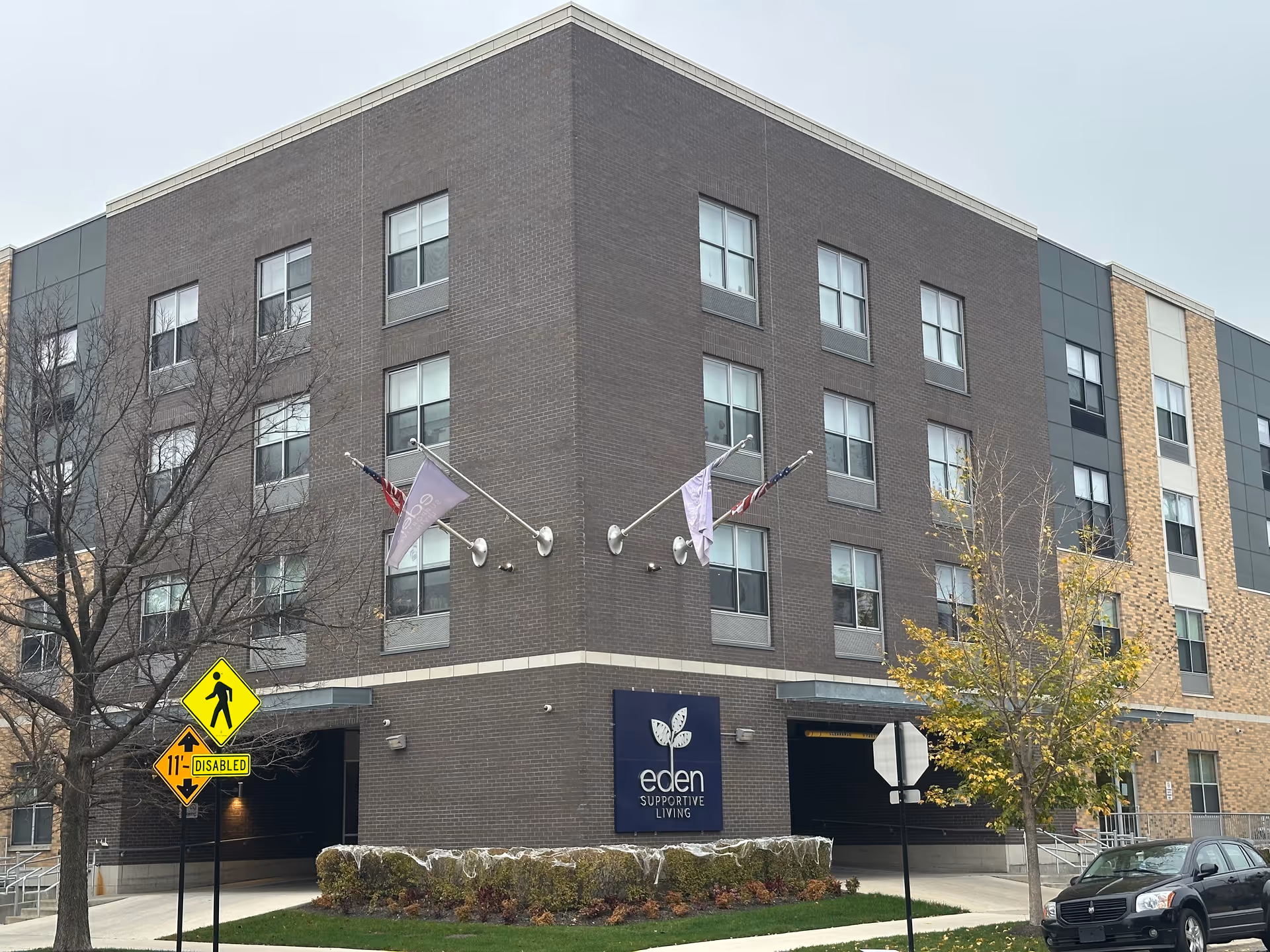 Exterior front of a multi-story brick building with an 'eden supportive living' sign and flags.