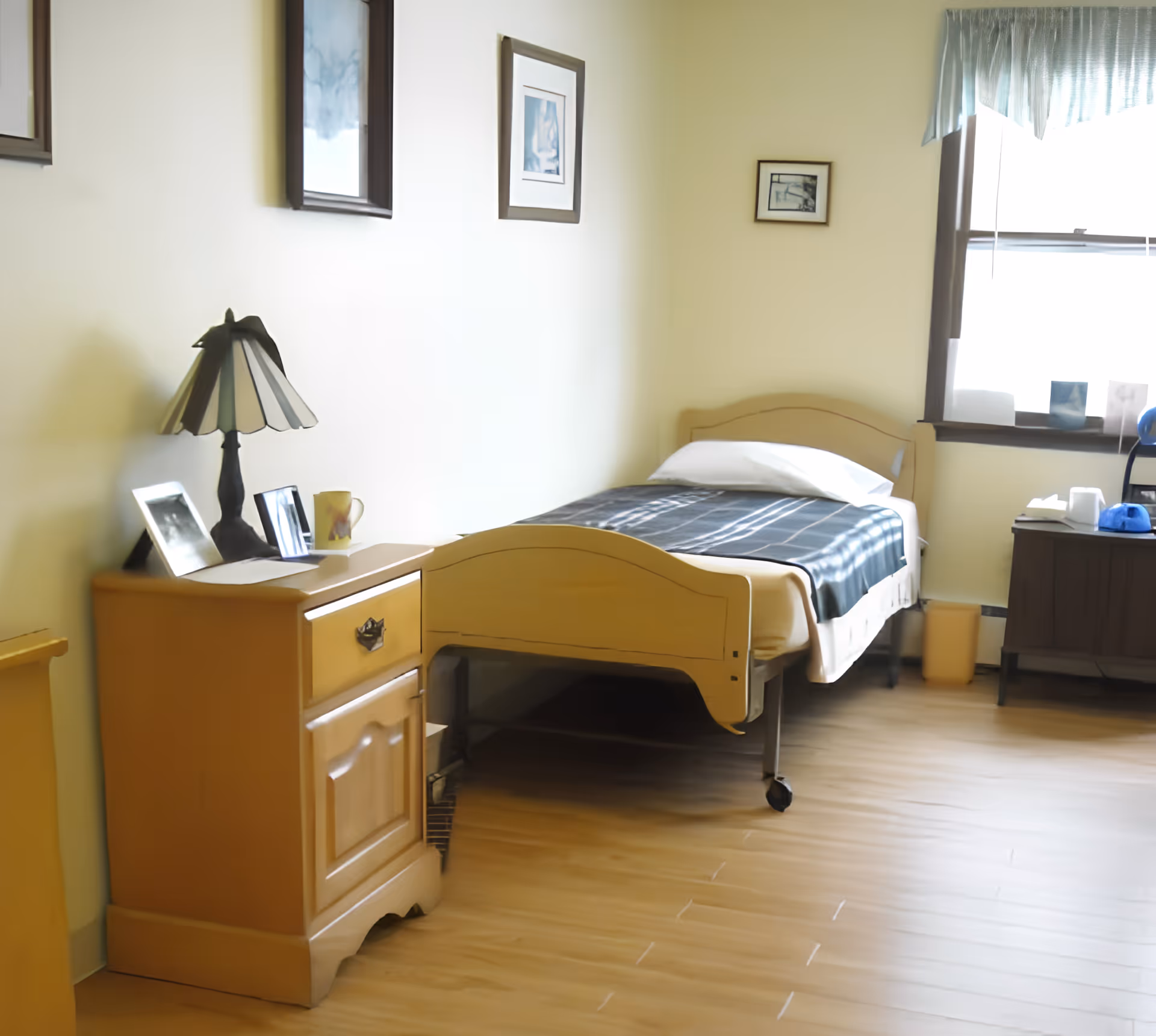 A simple bedroom in a senior living facility with a single bed covered with a dark plaid blanket and a white pillow. Next to the bed is a wooden nightstand with a lamp, framed photos, and a mug. The room has light-colored walls with three framed pictures hanging and a window with light blue curtains letting in natural light. There is a small cabinet near the window with some items on top.