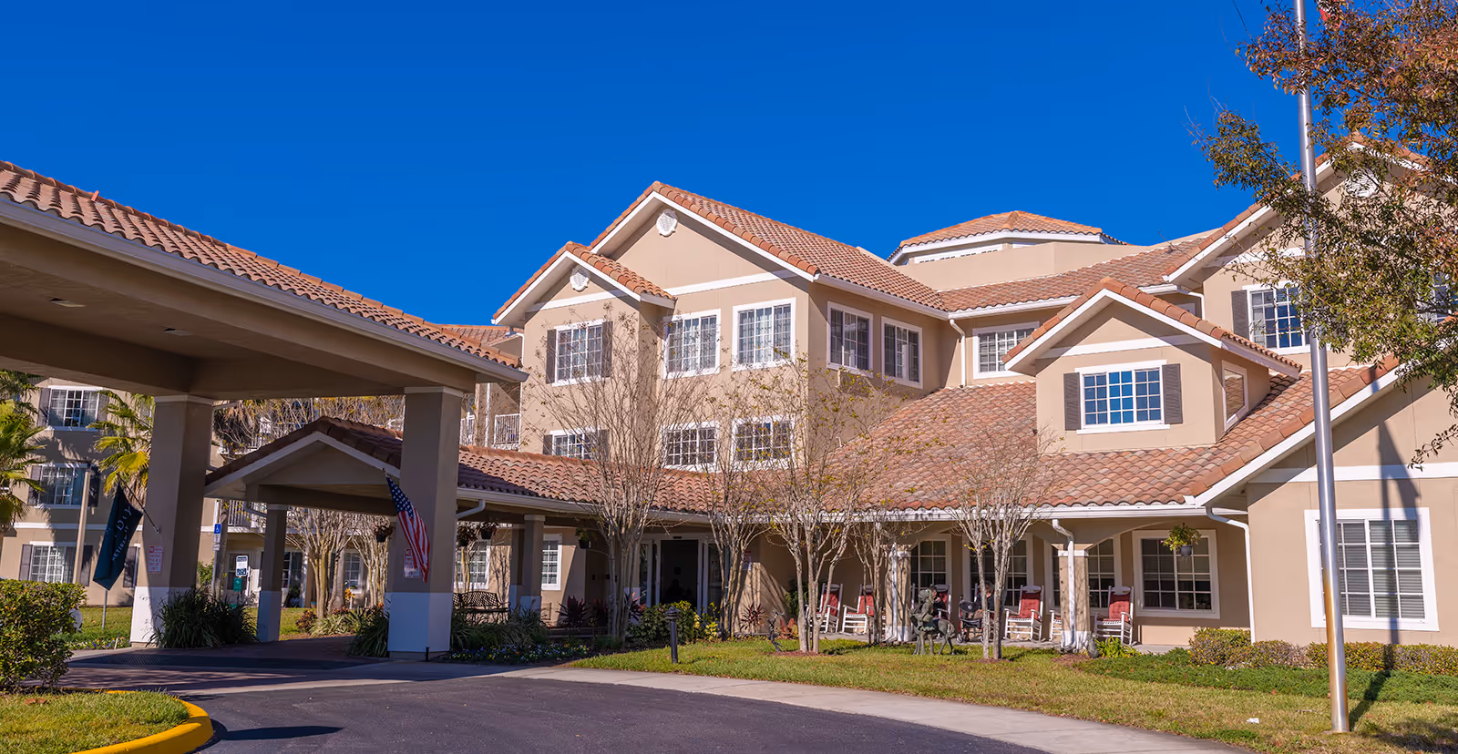 Exterior view of a senior living facility building with beige walls and red-tiled roofs under a clear blue sky. The entrance has a covered driveway with columns, and there are trees and shrubs around the building. Several rocking chairs are placed on the porch area.
