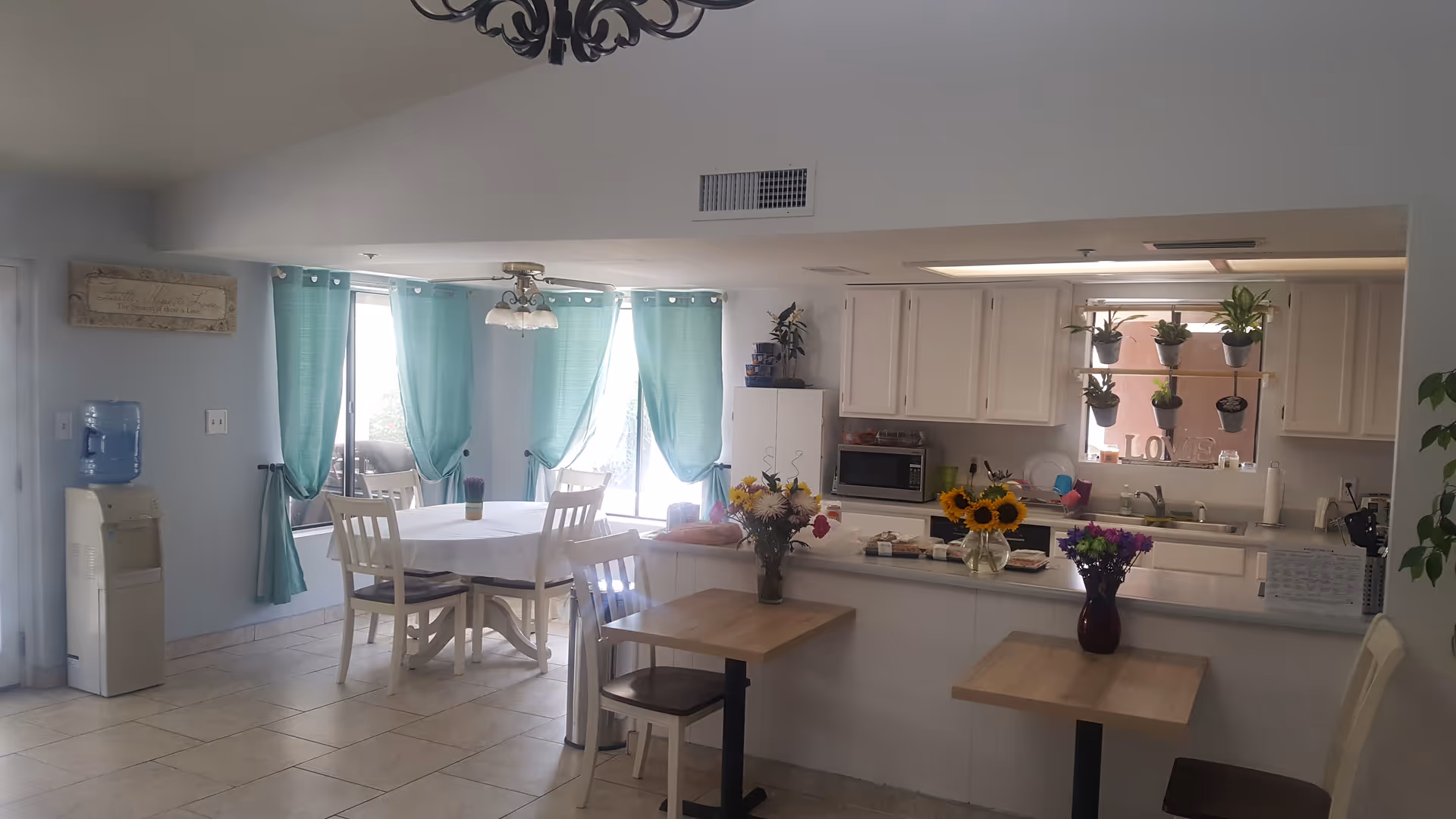 Bright dining area with turquoise curtains, white tables, and a visible kitchen in the background.