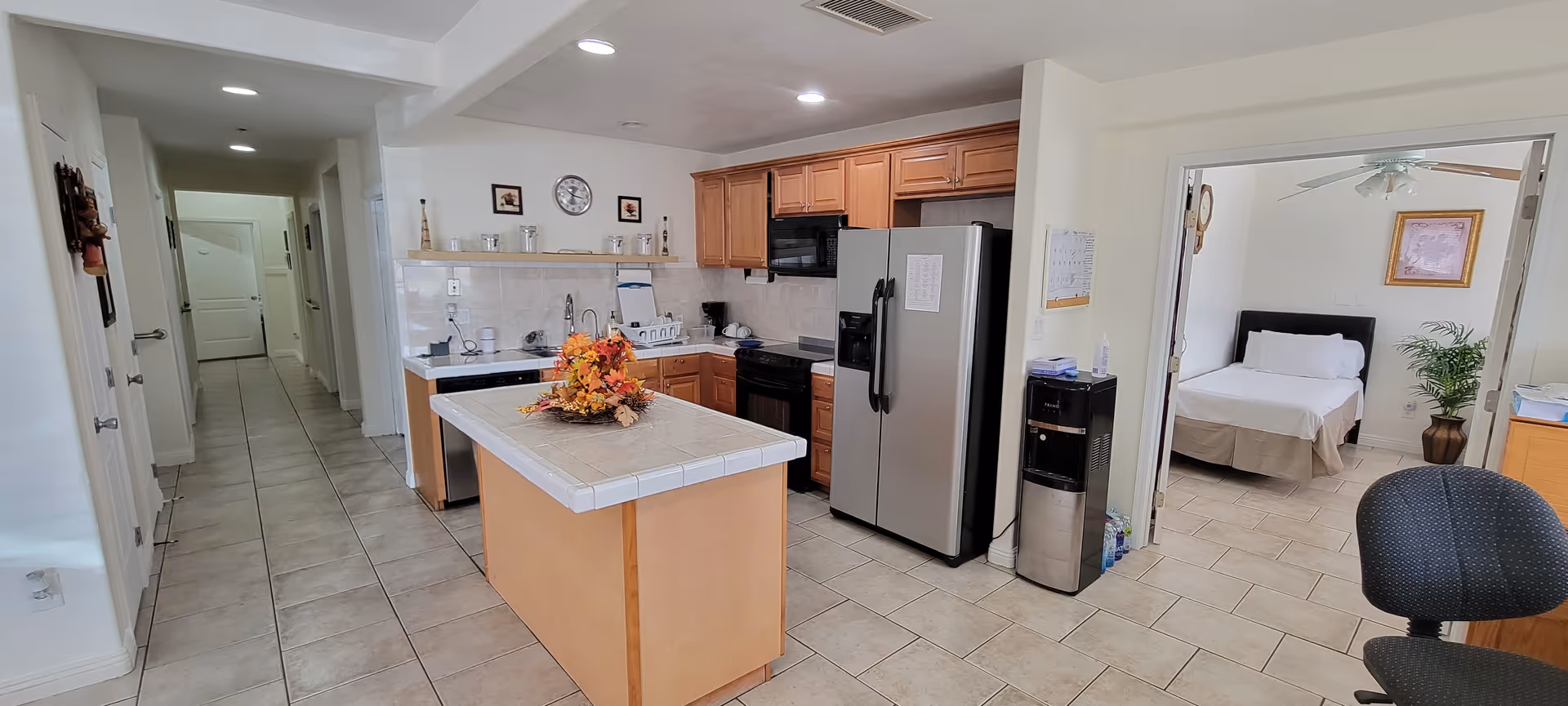 Open-concept kitchen with island, stainless fridge, and doorway to a bedroom in a senior facility.