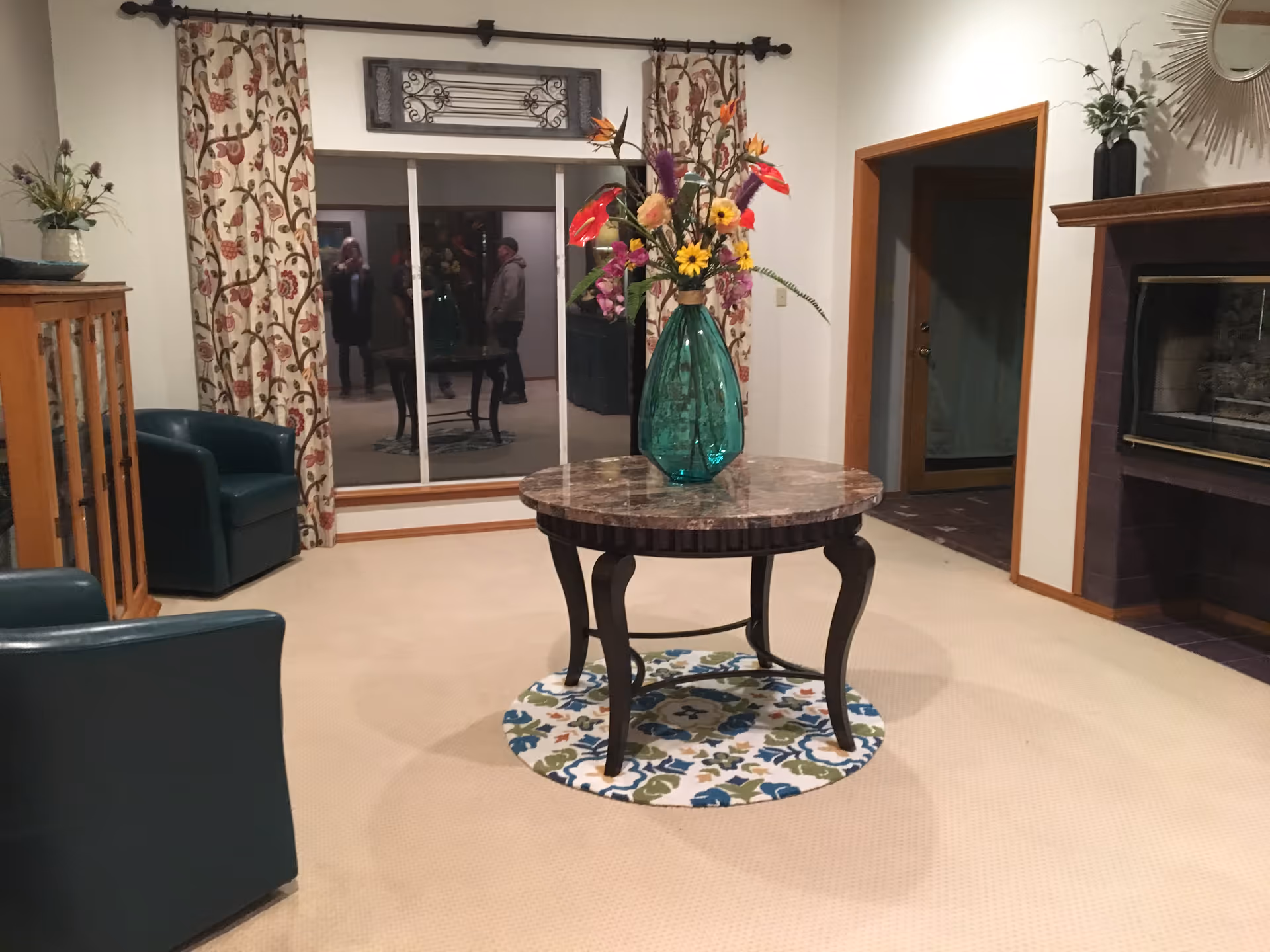 Interior lounge with round marble table, vibrant flower arrangement, green chairs, and a fireplace.
