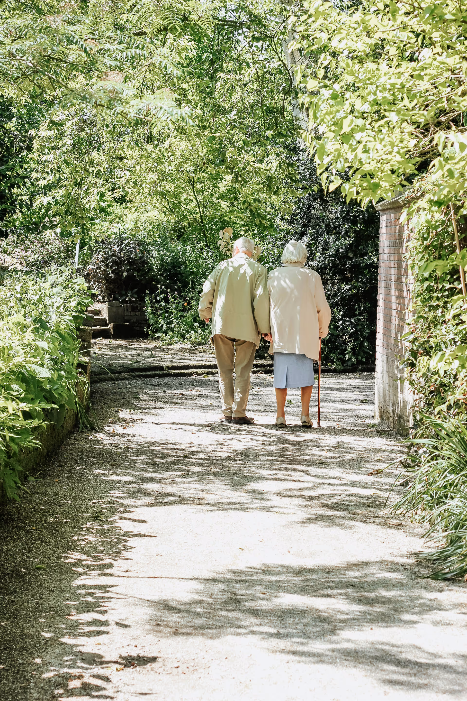 An elderly couple walking hand in hand down a sunlit garden path surrounded by lush green trees and plants. The woman is using a cane for support.