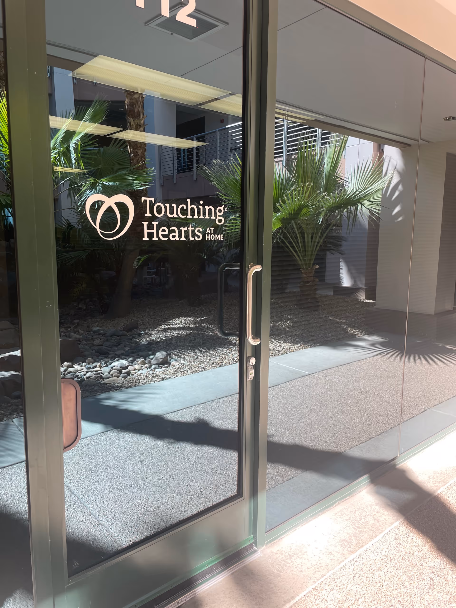 Glass entrance door with Touching Hearts at Home logo and surrounding plants.