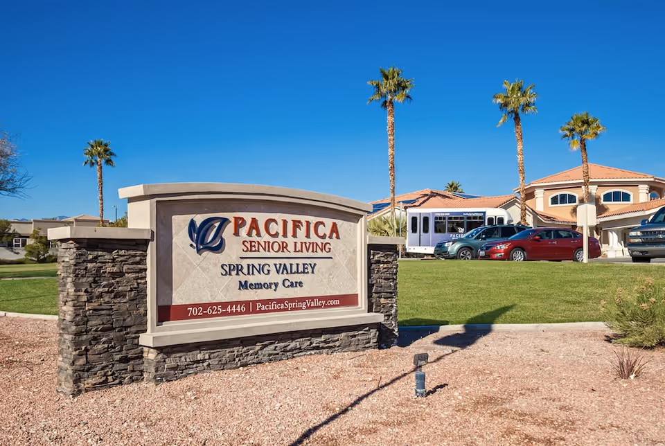 Entrance sign for Pacifica Senior Living Spring Valley Memory Care in front of the facility with palm trees and parked cars.