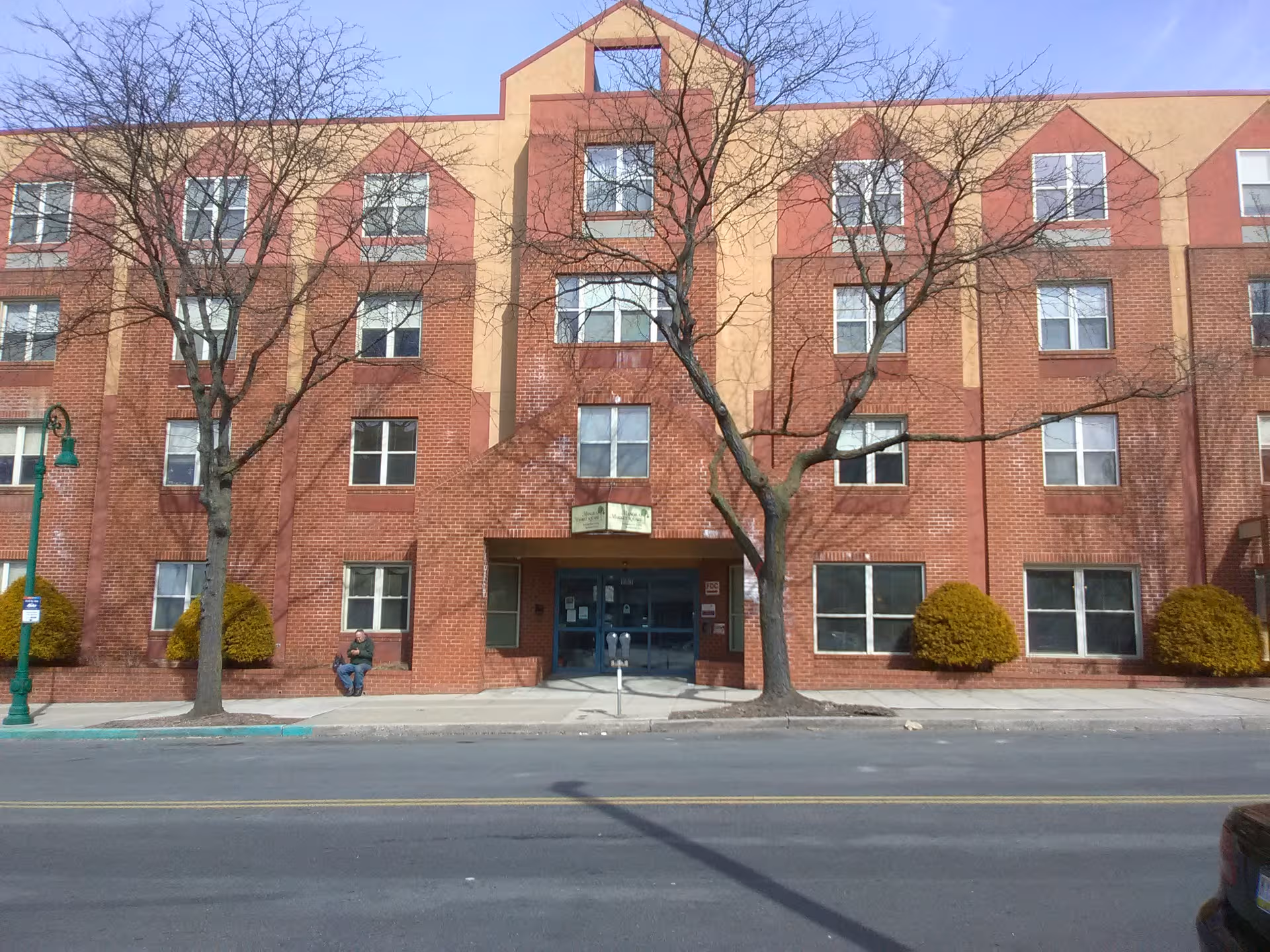 Red brick senior living building with trees and a sidewalk entrance along a city street