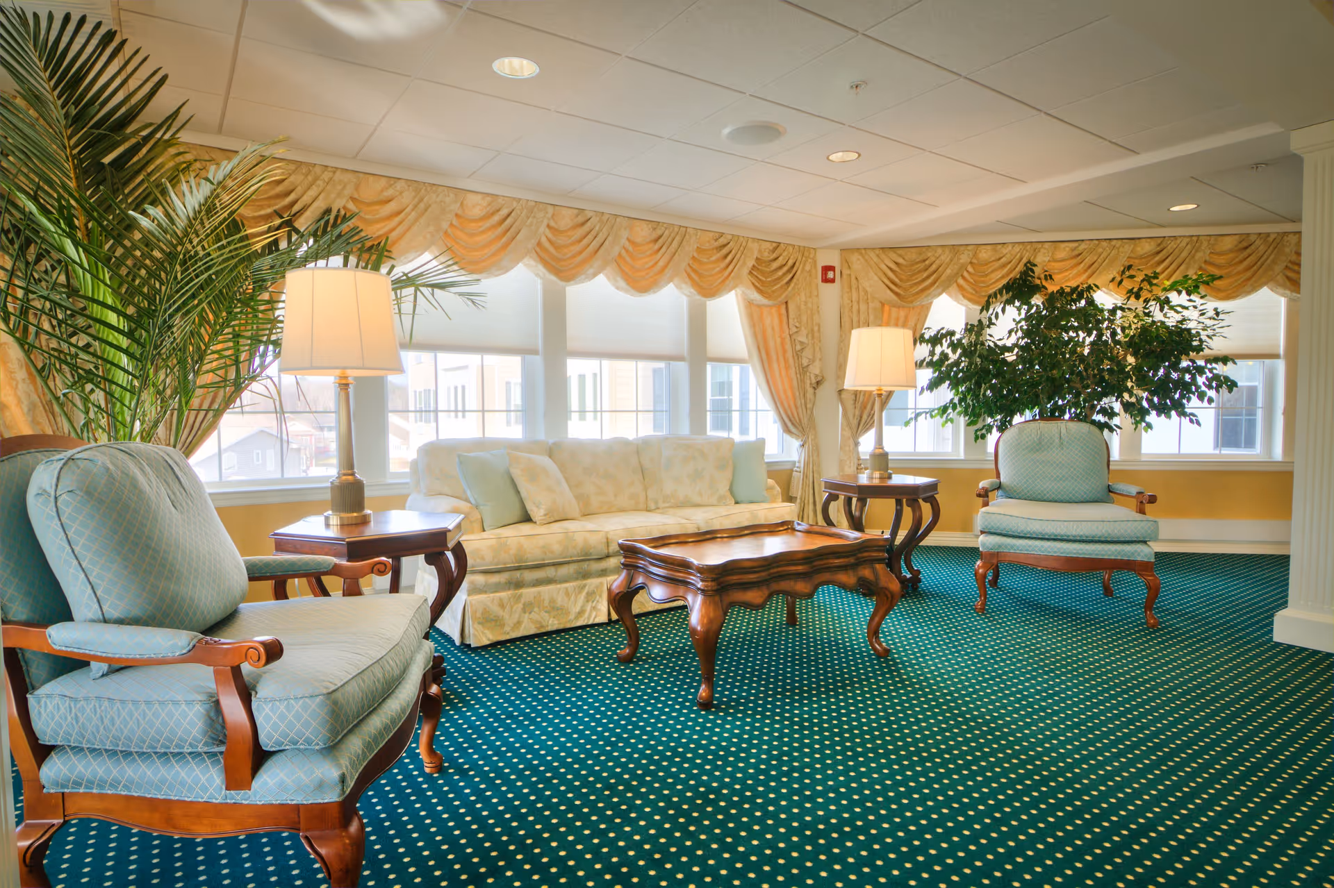 A bright and elegant living room with a green carpet featuring yellow dots, two light blue upholstered armchairs with wooden frames, a cream-colored sofa with patterned cushions, two wooden side tables each with a lamp, a wooden coffee table, large windows with yellow draped curtains, and green plants in the corners.