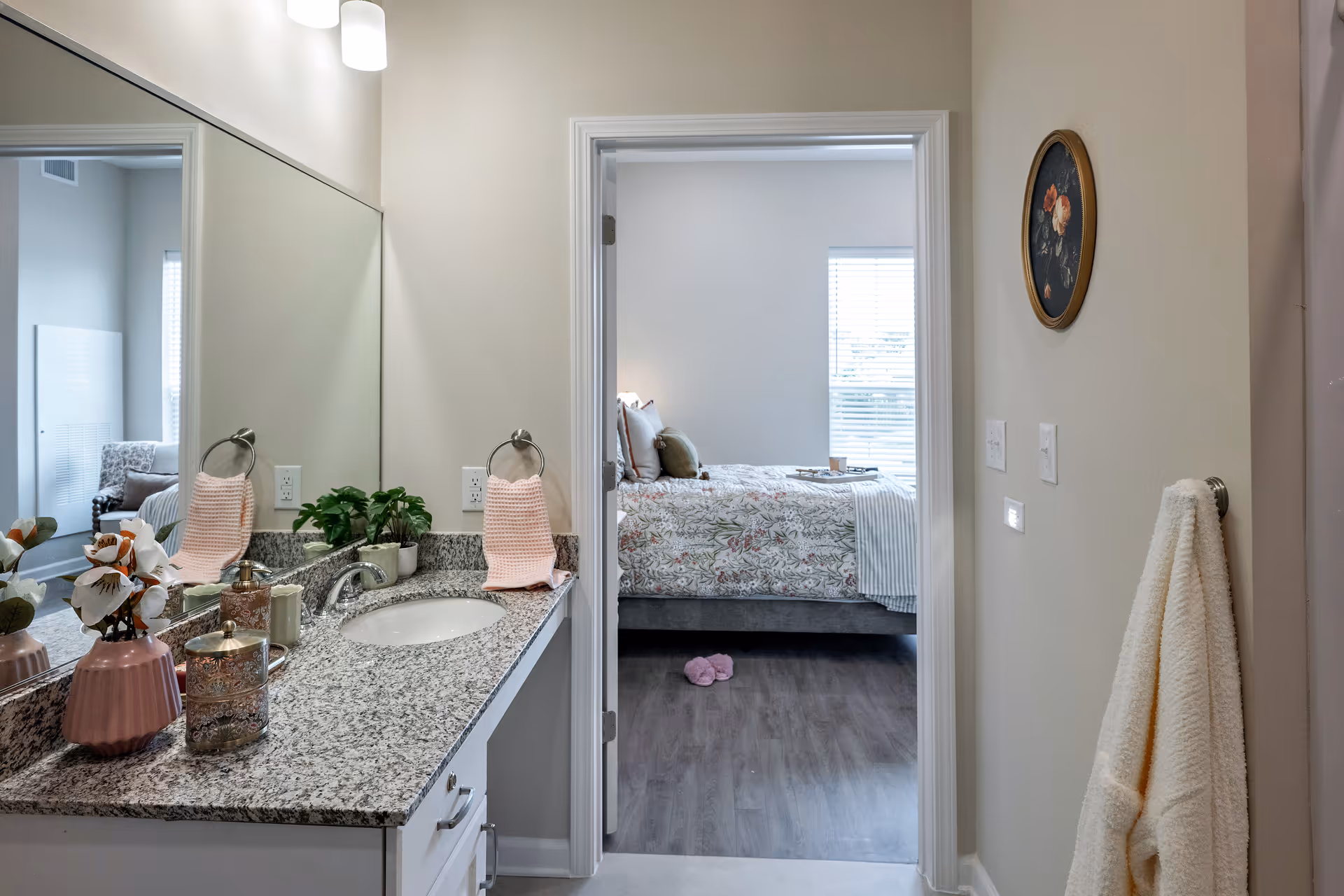 Bathroom vanity with granite countertop and mirror, opening to a bedroom with floral bedding.
