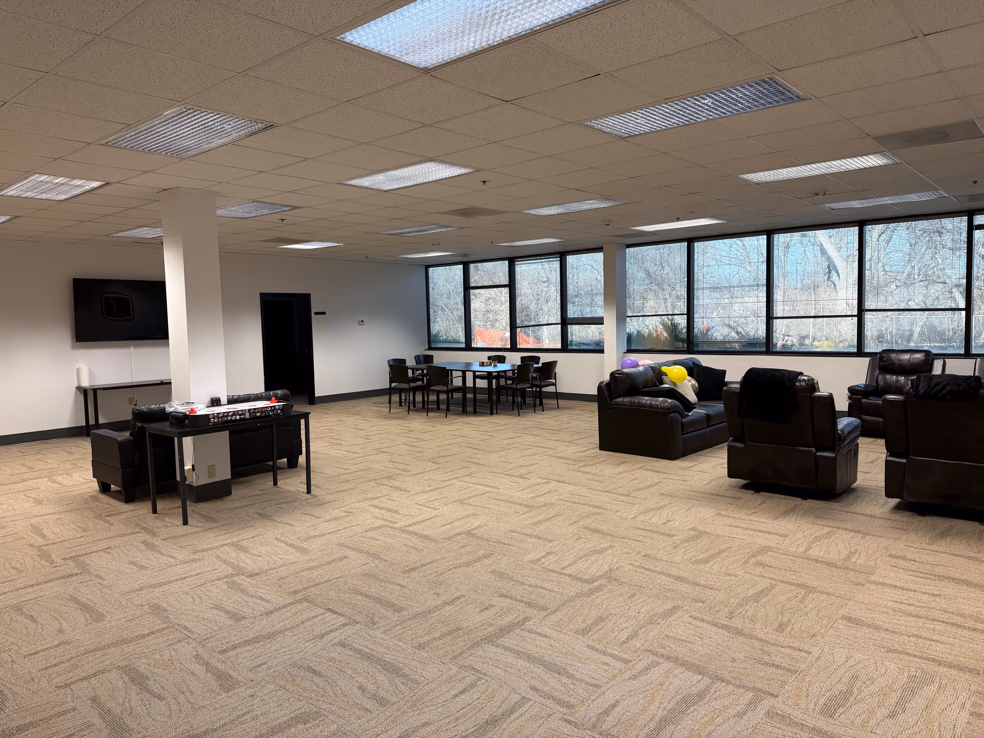 Spacious senior living lounge with brown leather seating, a foosball table, balloons, a TV, and windows.