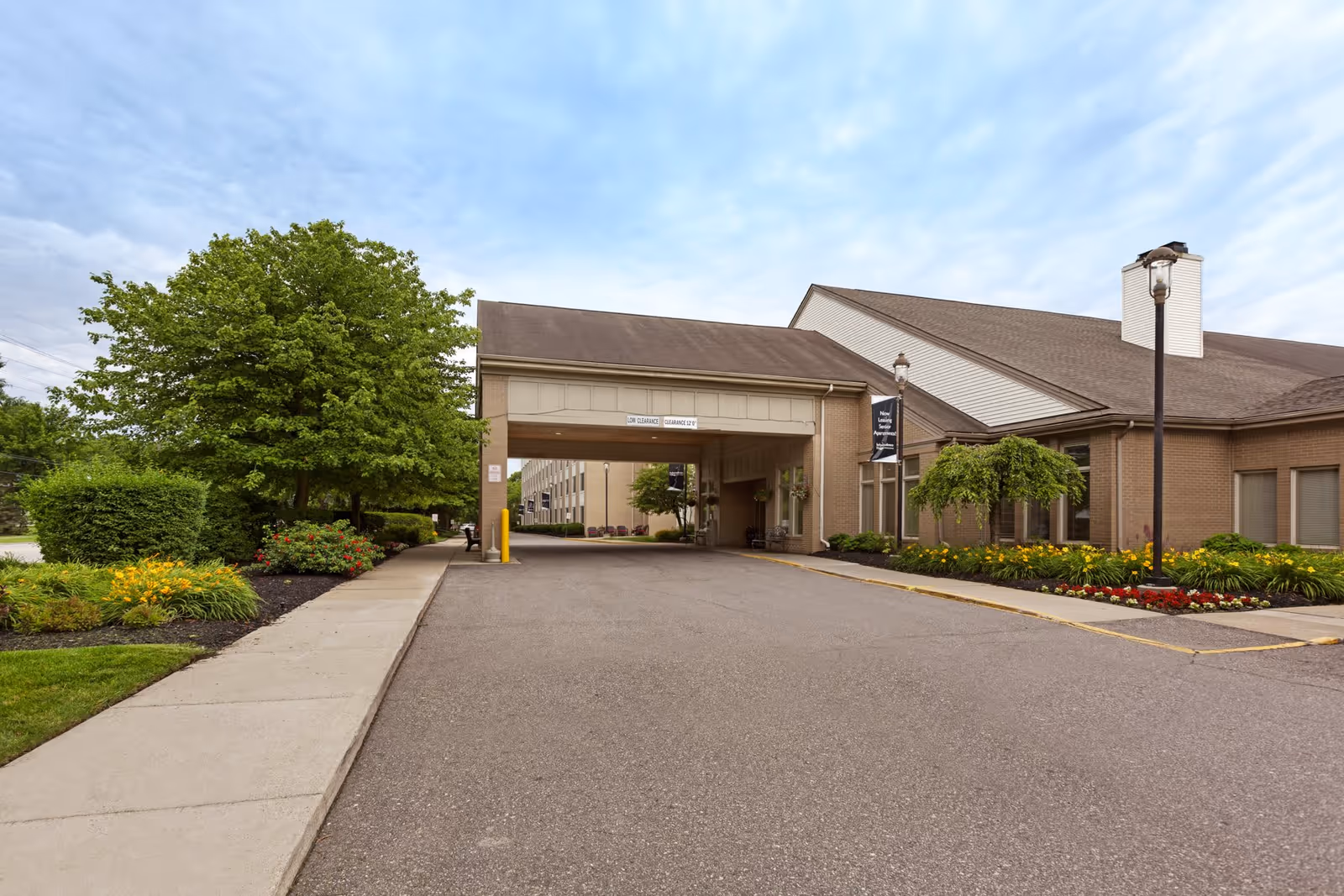 Entrance driveway to a senior living facility with a covered drop-off area. The building is made of brick with a sloped roof. There are well-maintained flower beds and green trees on both sides of the driveway under a partly cloudy sky.