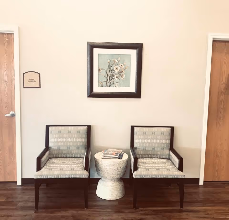 Two patterned armchairs with wooden frames are placed on either side of a small white round table holding magazines. Above the table hangs a framed floral painting. There are two wooden doors on either side of the chairs, with a sign on the left door that reads 'Social Services'. The floor is wooden and the walls are light-colored.