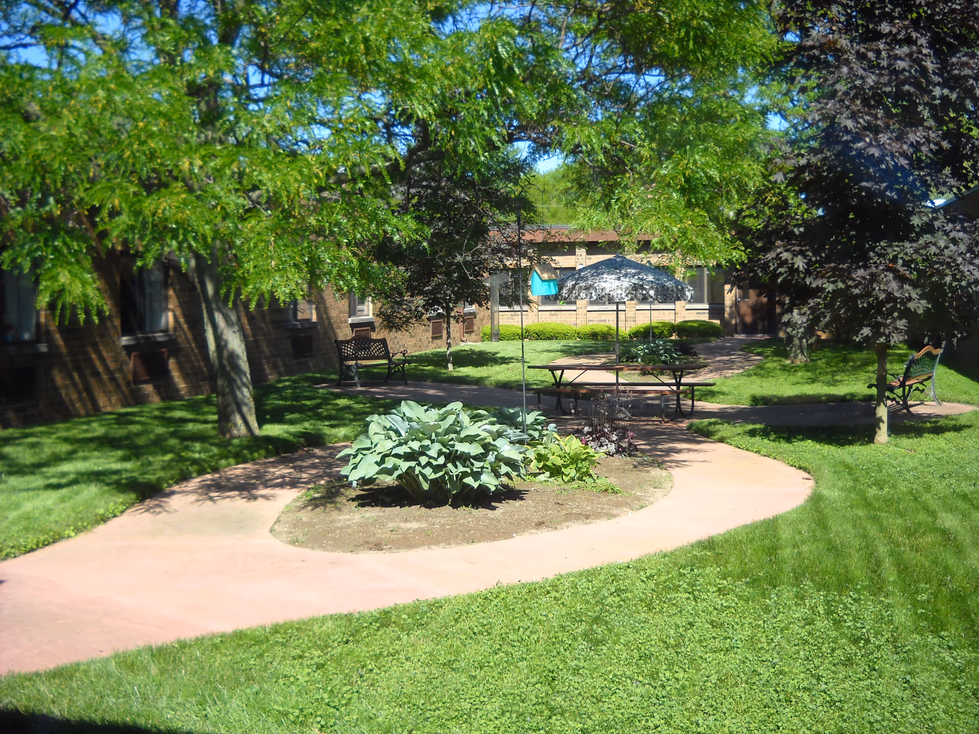 A sunny outdoor courtyard with green grass, trees, and a curved concrete walkway. There are benches along the path and a picnic table with an umbrella in the center. The courtyard is surrounded by a brick building.