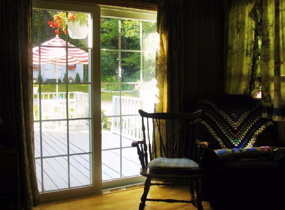 View from inside a dimly lit room showing a wooden chair with a cushion and an armchair draped with a colorful crocheted blanket. Through the large sliding glass door, a bright outdoor patio with white chairs and a red and white striped umbrella is visible, along with green trees and a white shed in the background.
