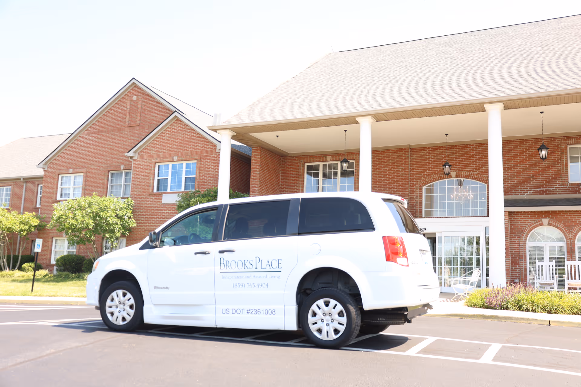 A white van parked in front of a brick senior living building with columns at the covered entrance.