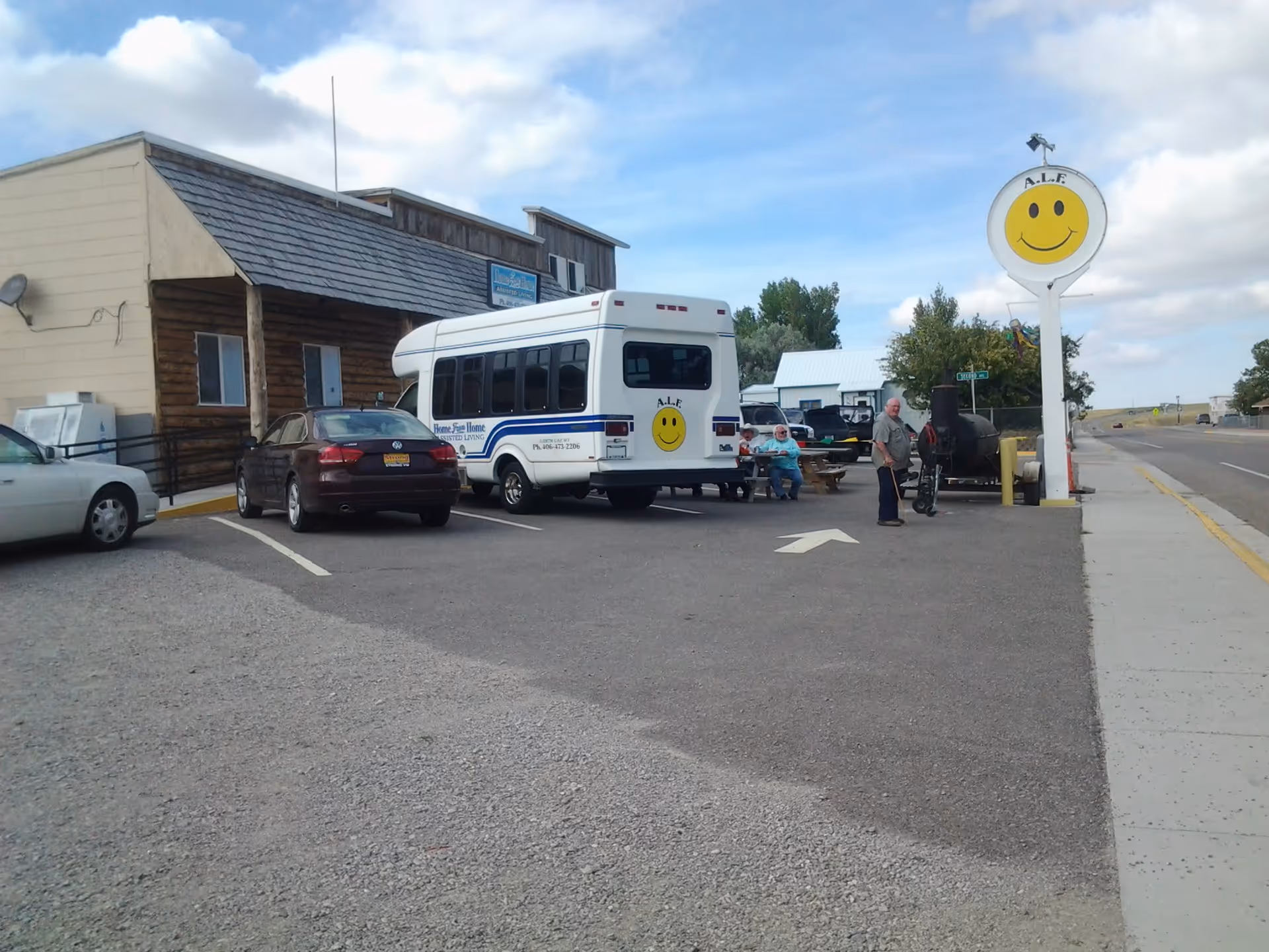 Parking lot outside a building with a wooden facade and a signpost featuring a large yellow smiley face and the letters A.L.F. Several vehicles, including a white shuttle bus with a smiley face on the back, are parked. Two people are sitting on a bench near the bus, and one person is standing nearby. The sky is partly cloudy.