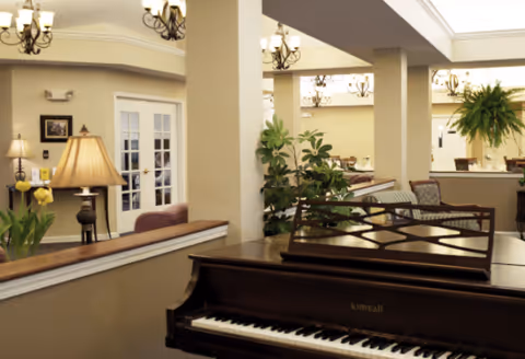 Interior view of a senior living facility lounge area featuring a Kimball piano in the foreground, decorative plants, a lamp on a side table, framed pictures on the wall, and multiple chandeliers hanging from the ceiling. The space has beige walls and white trim with a cozy and inviting atmosphere.
