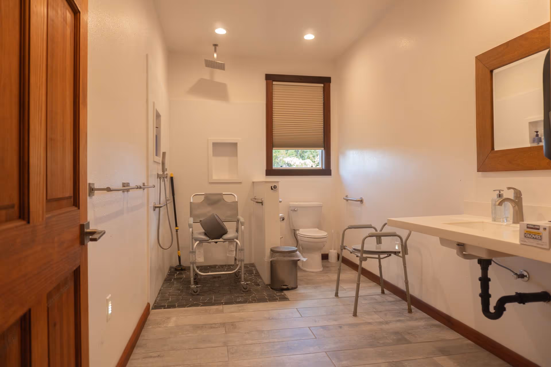 Accessible bathroom with ceiling shower, toilet, sink, chair, and grab bars.