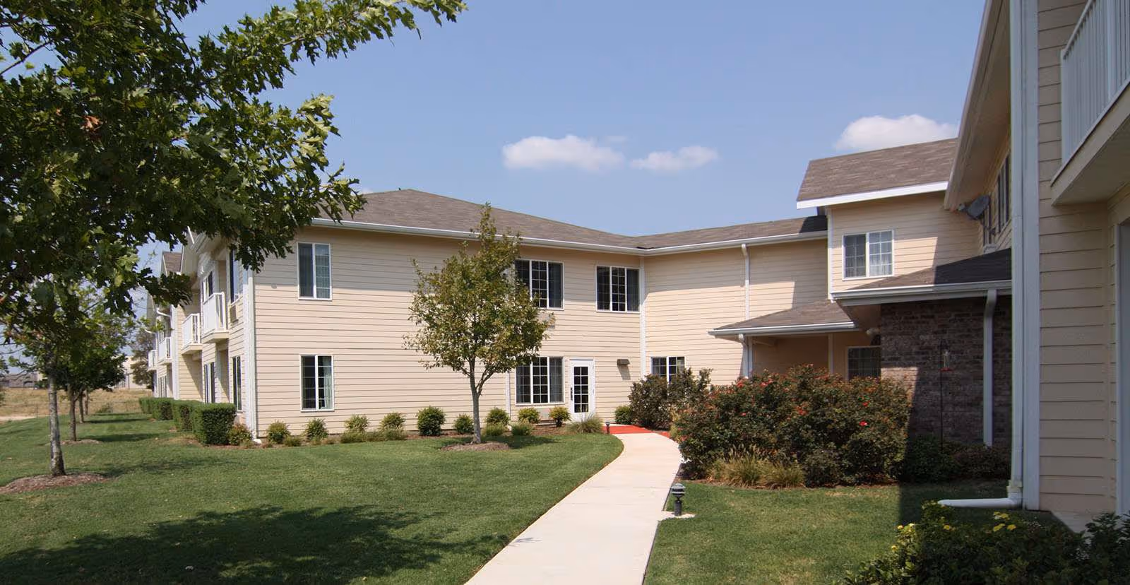 Walkway leading through a landscaped lawn to the front entrance of a two-story beige senior living building under a blue sky.